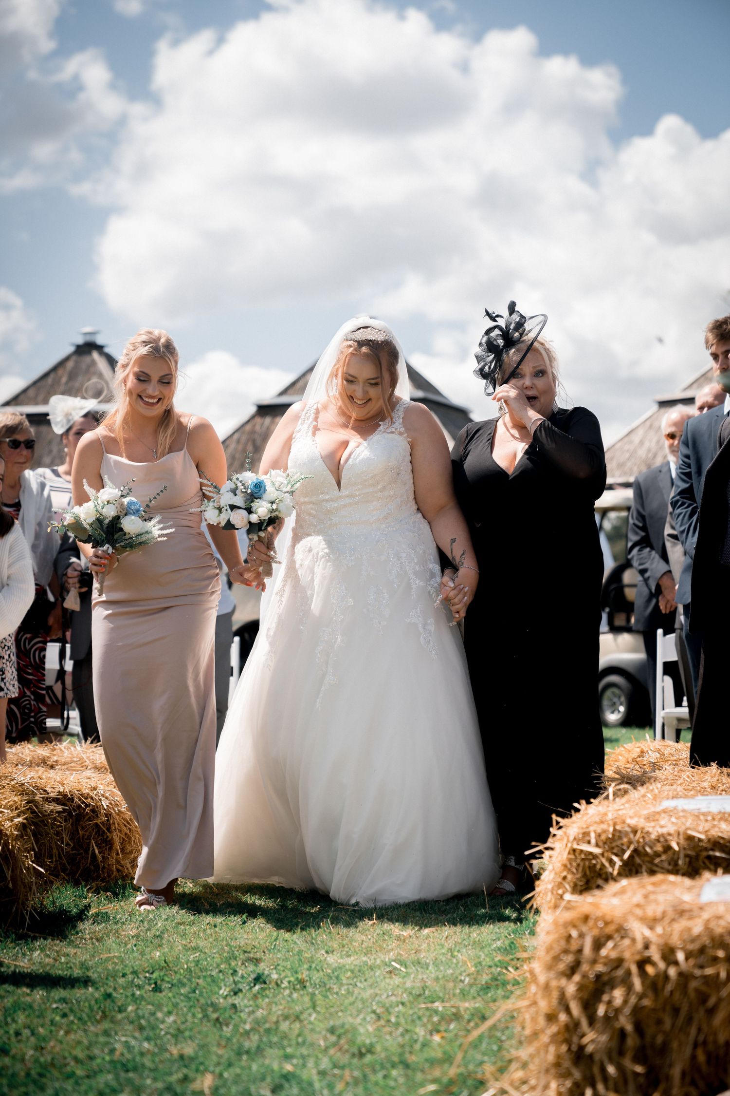 Bride walking down outdoor aisle with bridesmaids past hay bales at a rustic farm wedding ceremony.