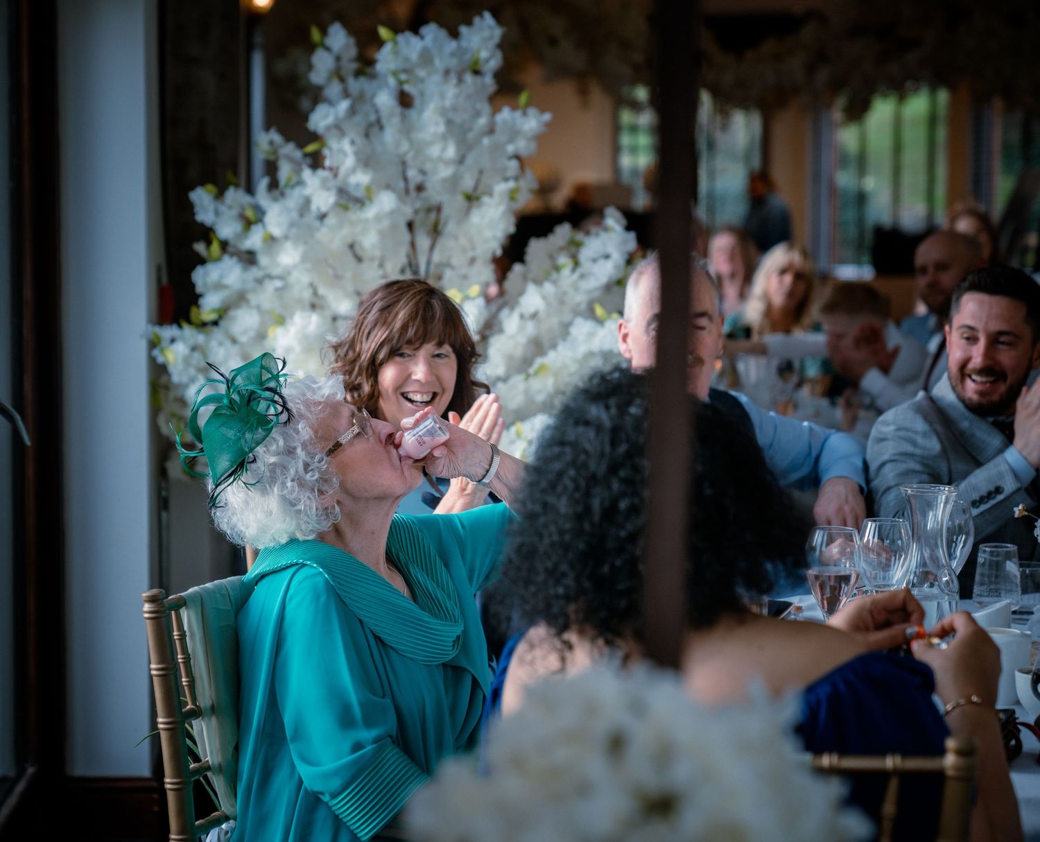 Wedding guests laughing and celebrating at a reception table adorned with white floral arrangements.