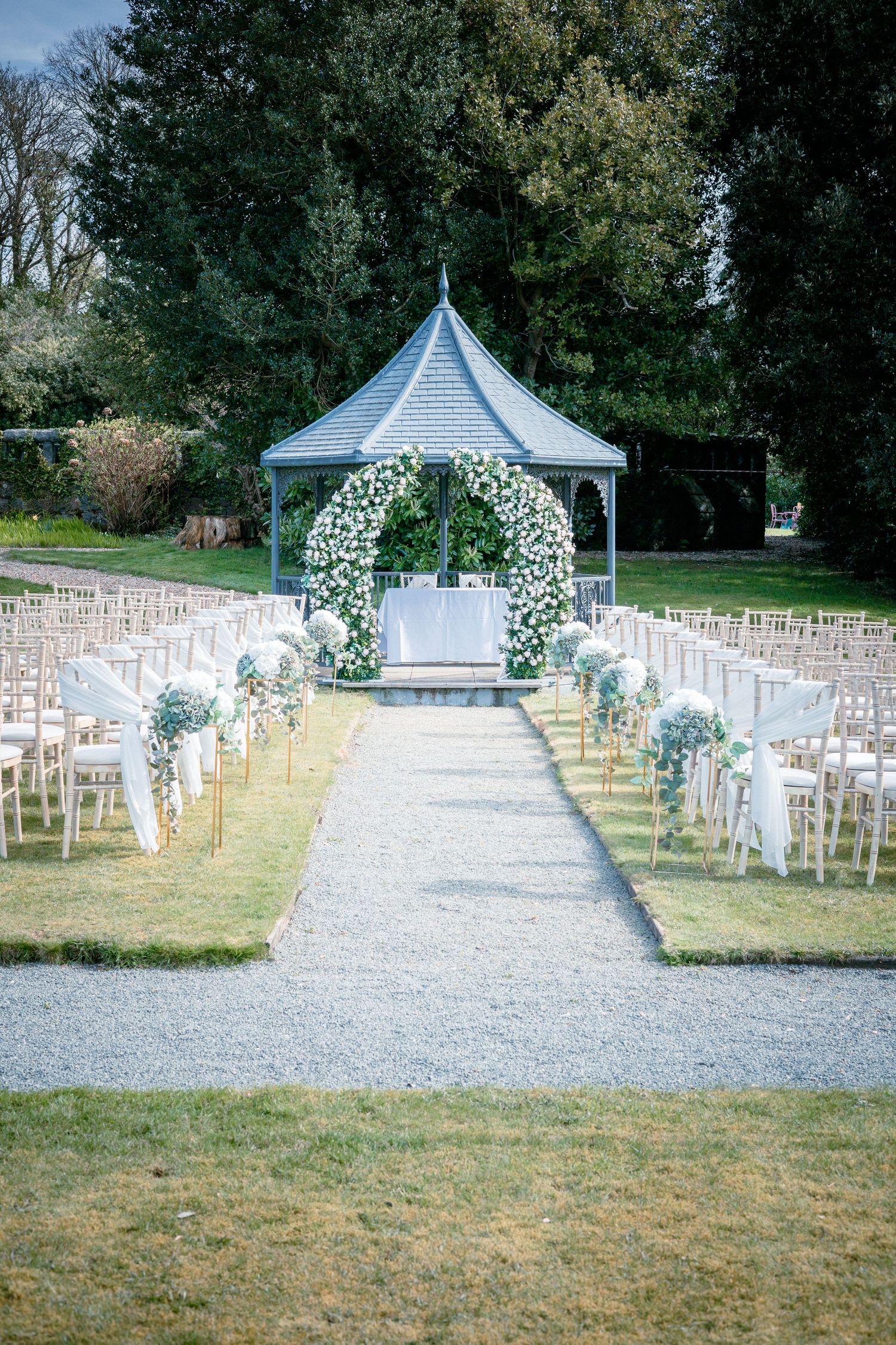 Outdoor wedding ceremony setup with floral arch, white chairs, and a gravel aisle in a garden.