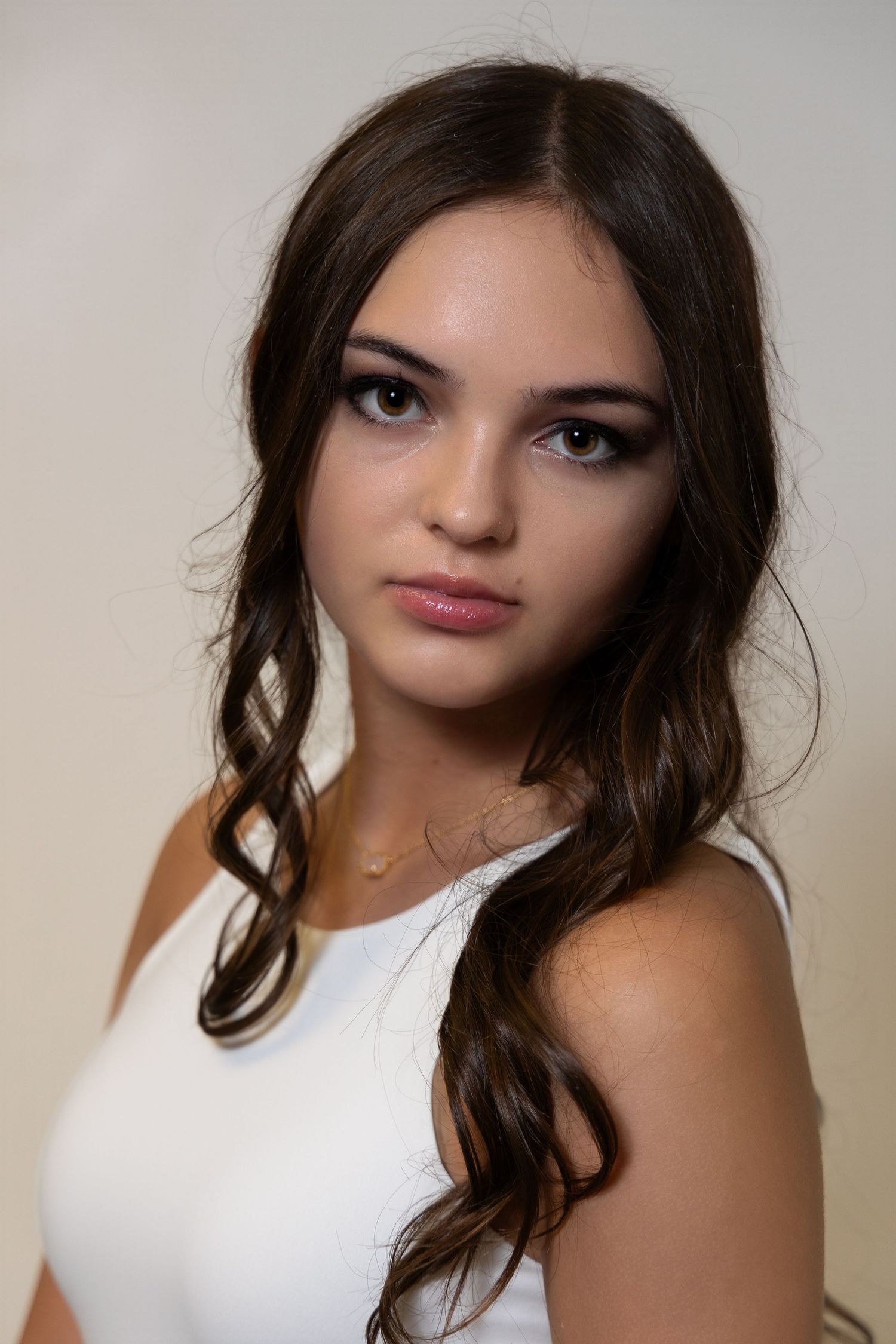 teenage girl wearing a white tank top with long brunette hair looking over her shoulder