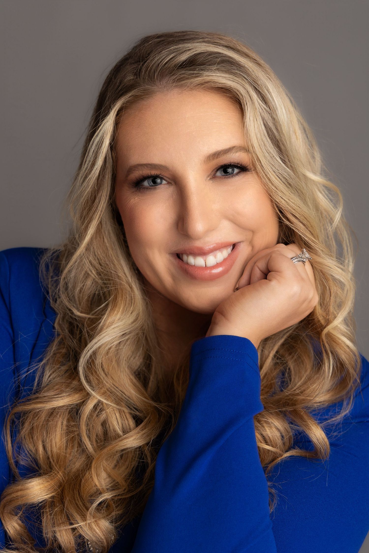 pageant headshot of blonde female with long curly hair wearing a blue blazer hand supporting chin