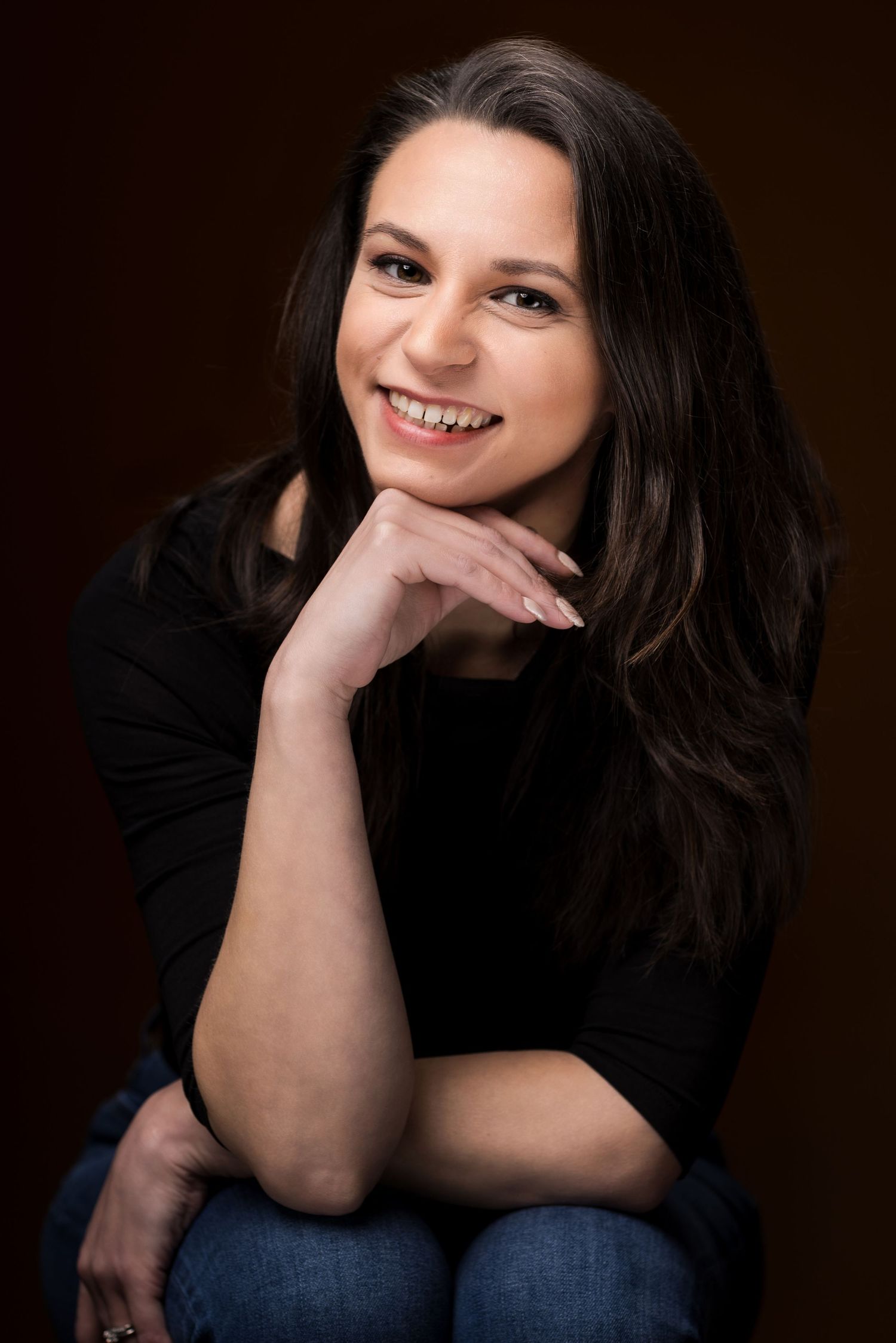 seated headshot of brunette female supporting chin with hand wearing black shirt and jeans