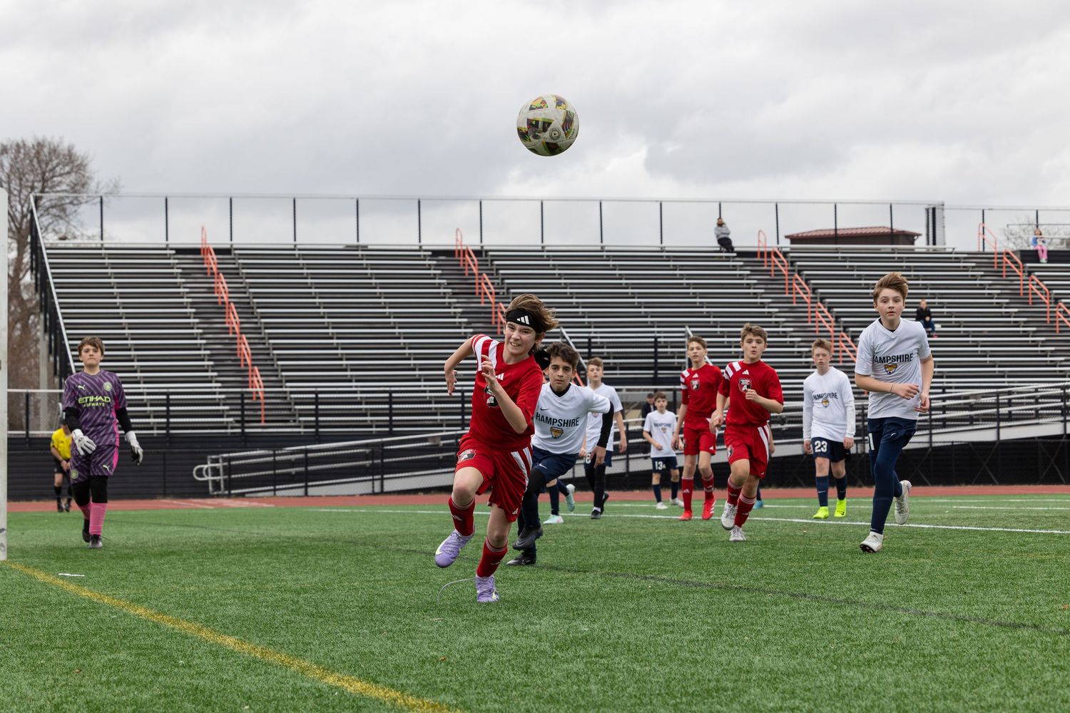 action sports game soccer group of teens male player wearing red, white uniforms running to a ball flying through air towards the camera