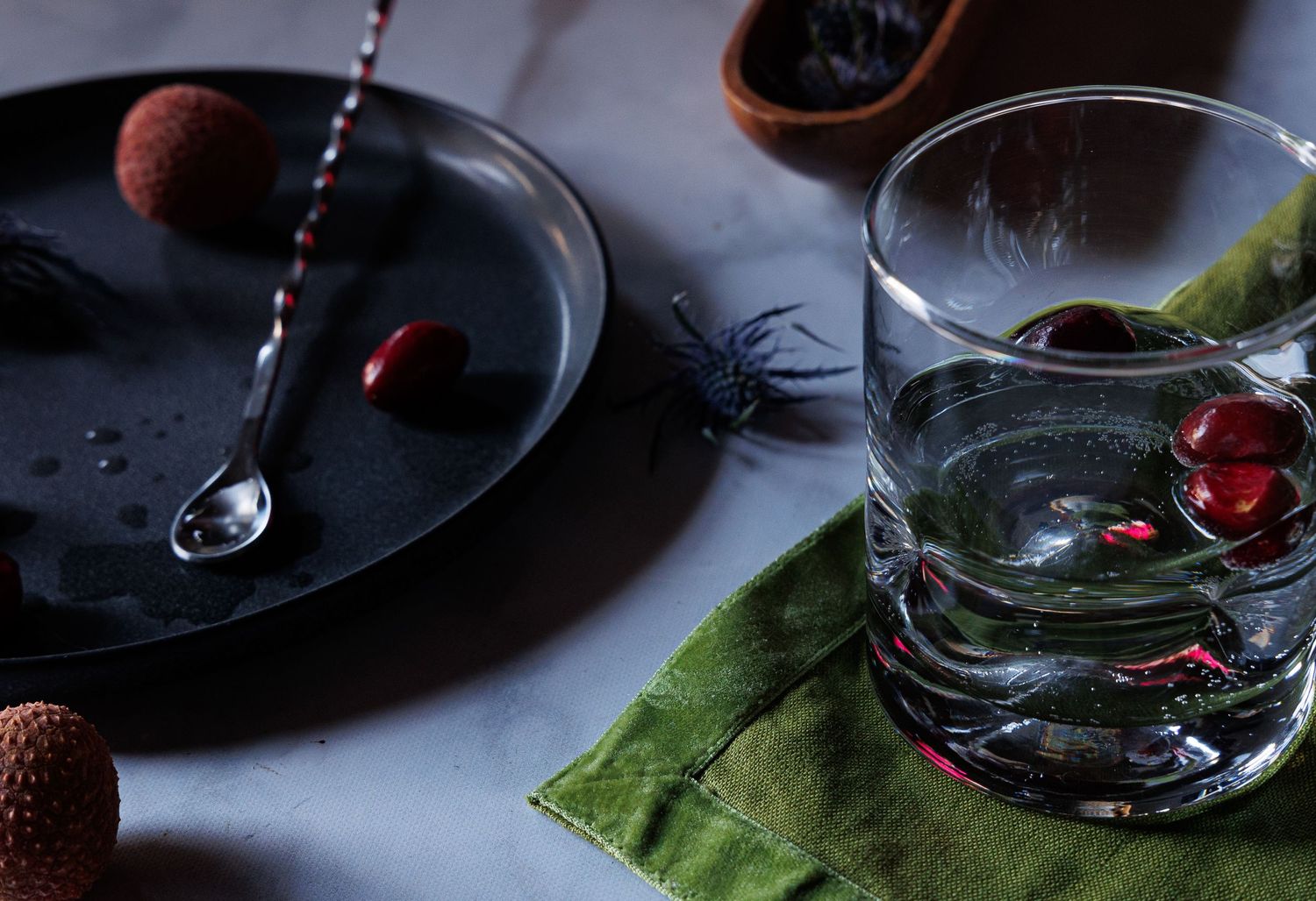 still life image of a table scape, black plate with drink spoon and fruit, white table cloth and green napkin, highball glass of water