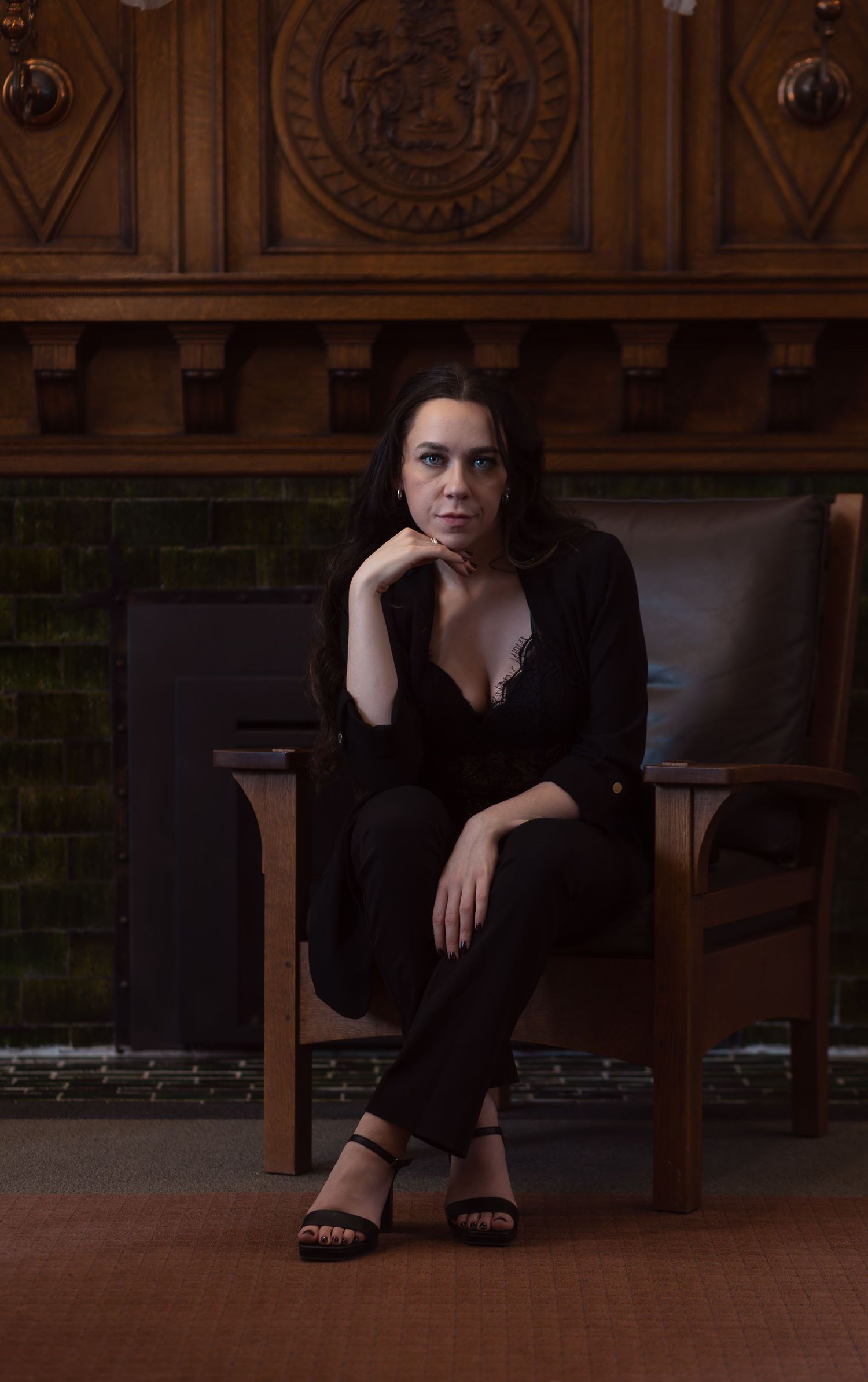 full body seated portrait of a brunette female sitting in a large wooden chair in front of an antique fireplace with her ankles crossed