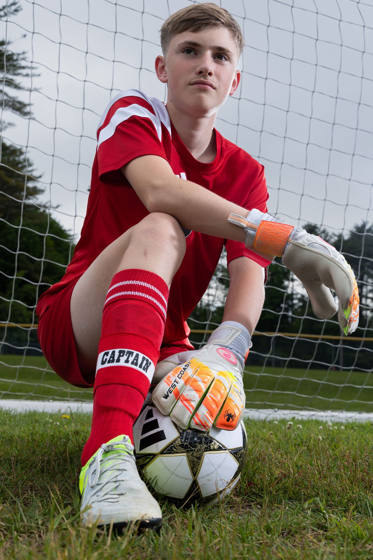 sports portrait blonde male teen soccer player goalkeeper kneeling in front of goal hand on ball wearing red uniform keeper gloves