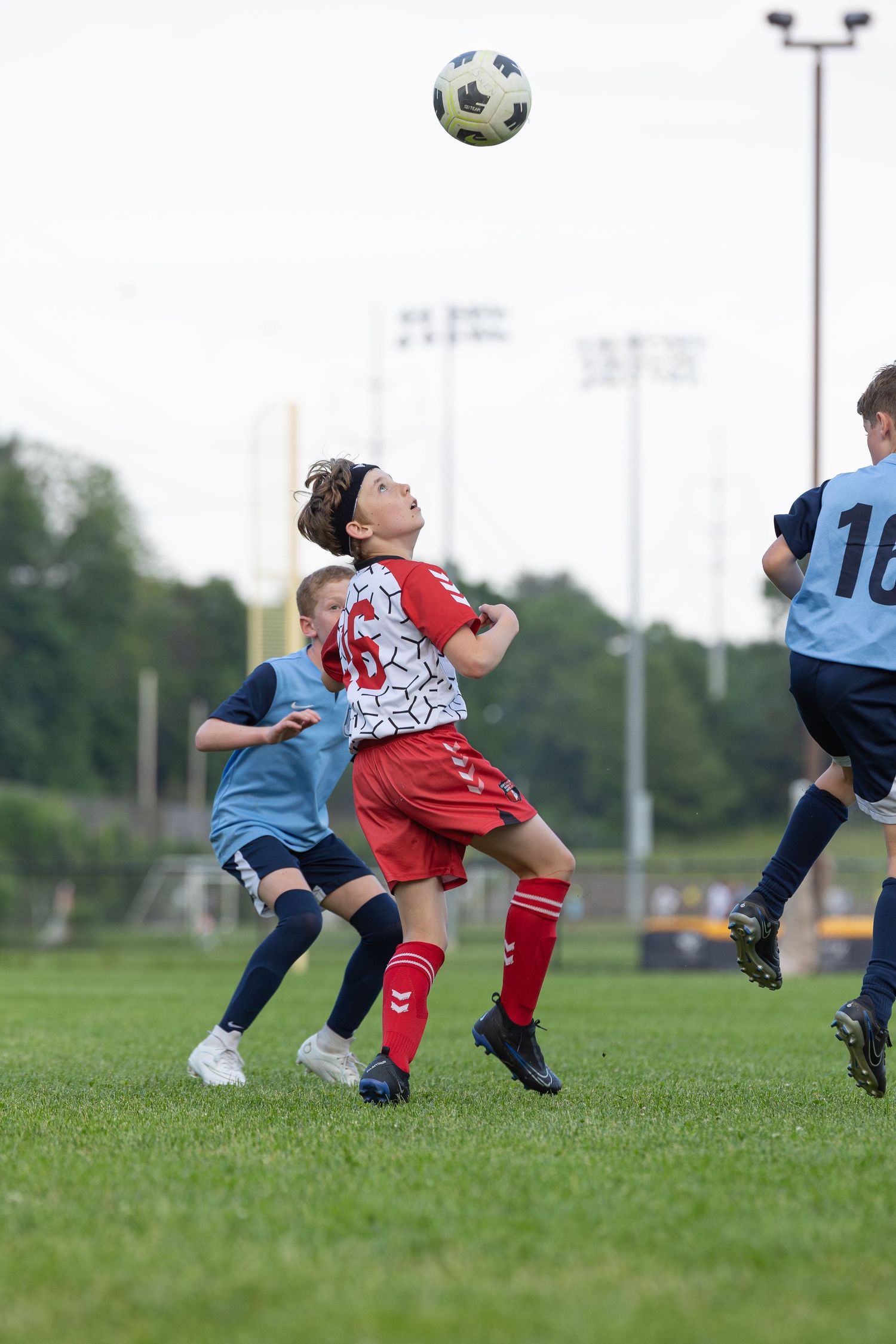 action sports game soccer teen male player red and white uniform looking up at soccer ball hanging in the air above head on a soccer field