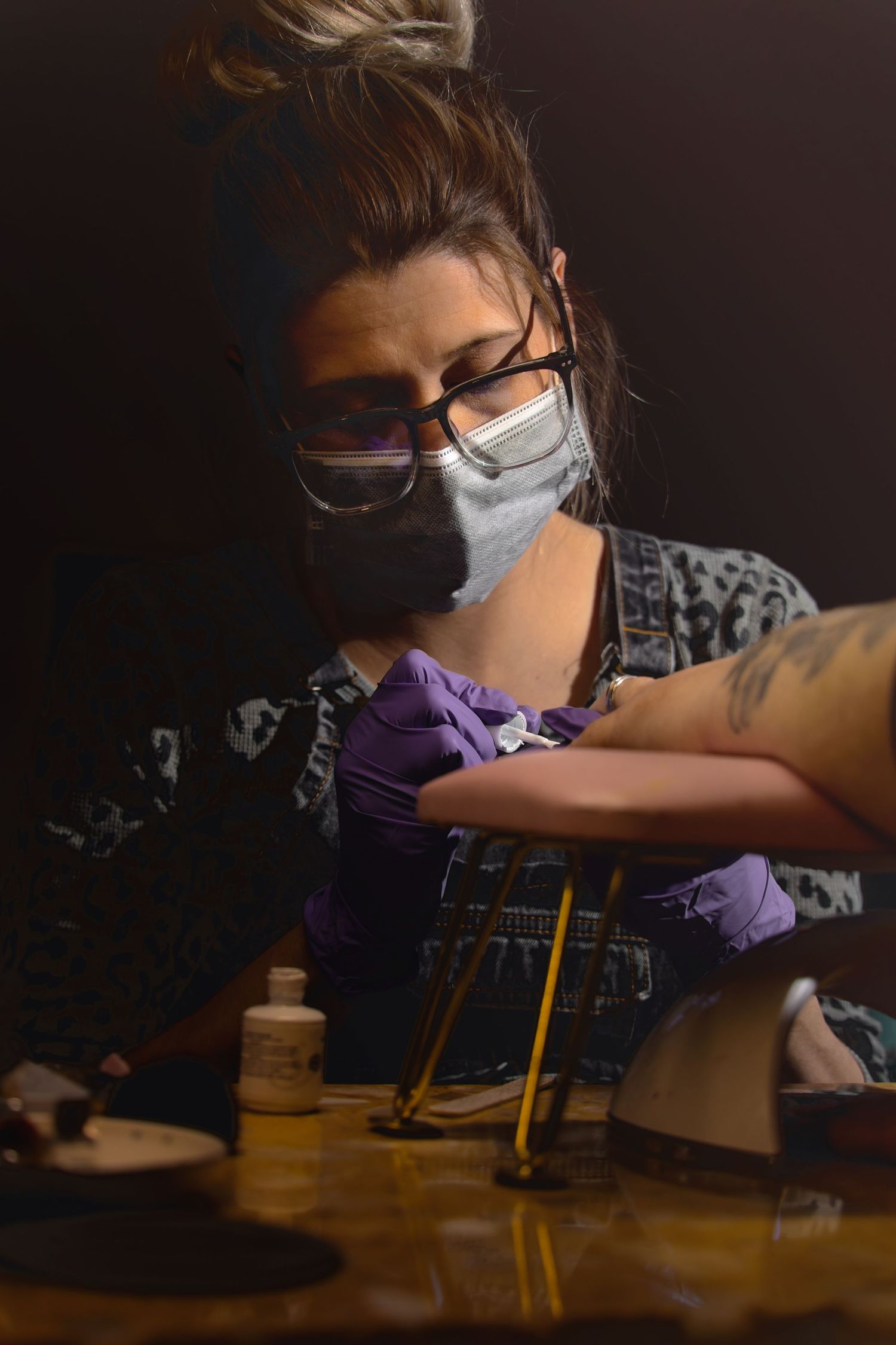 brand image of a blonde female nail tech wearing glasses painting the nails of a client whose hands are on a pink wrist rest