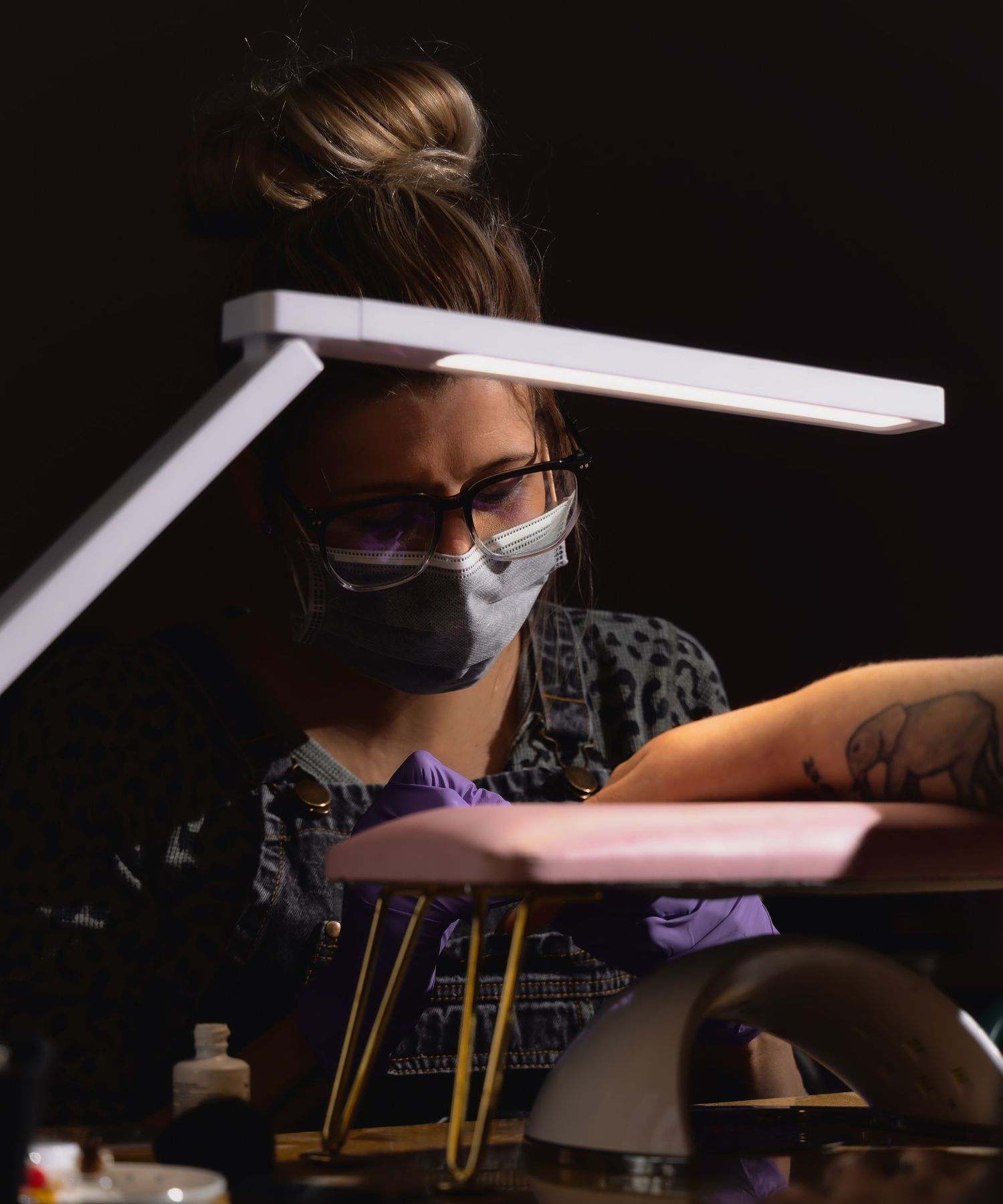 brand image of a blonde female nail tech wearing glasses painting the nails of a client whose hands are on a pink wrist rest