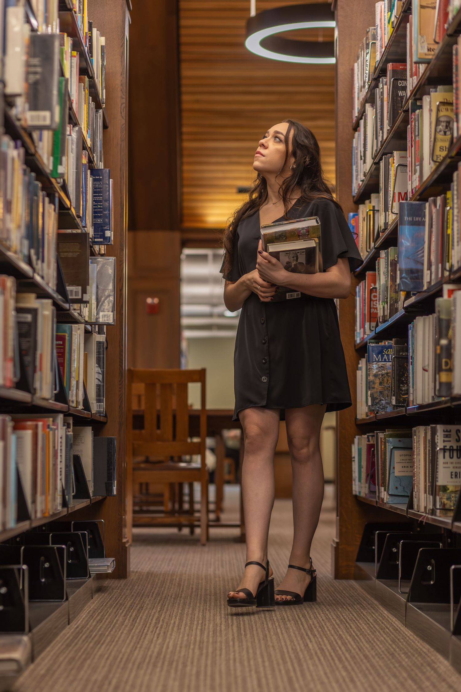 full body portrait of brunette woman wearing a black dress and heels walking down library aisle looking at and carrying books