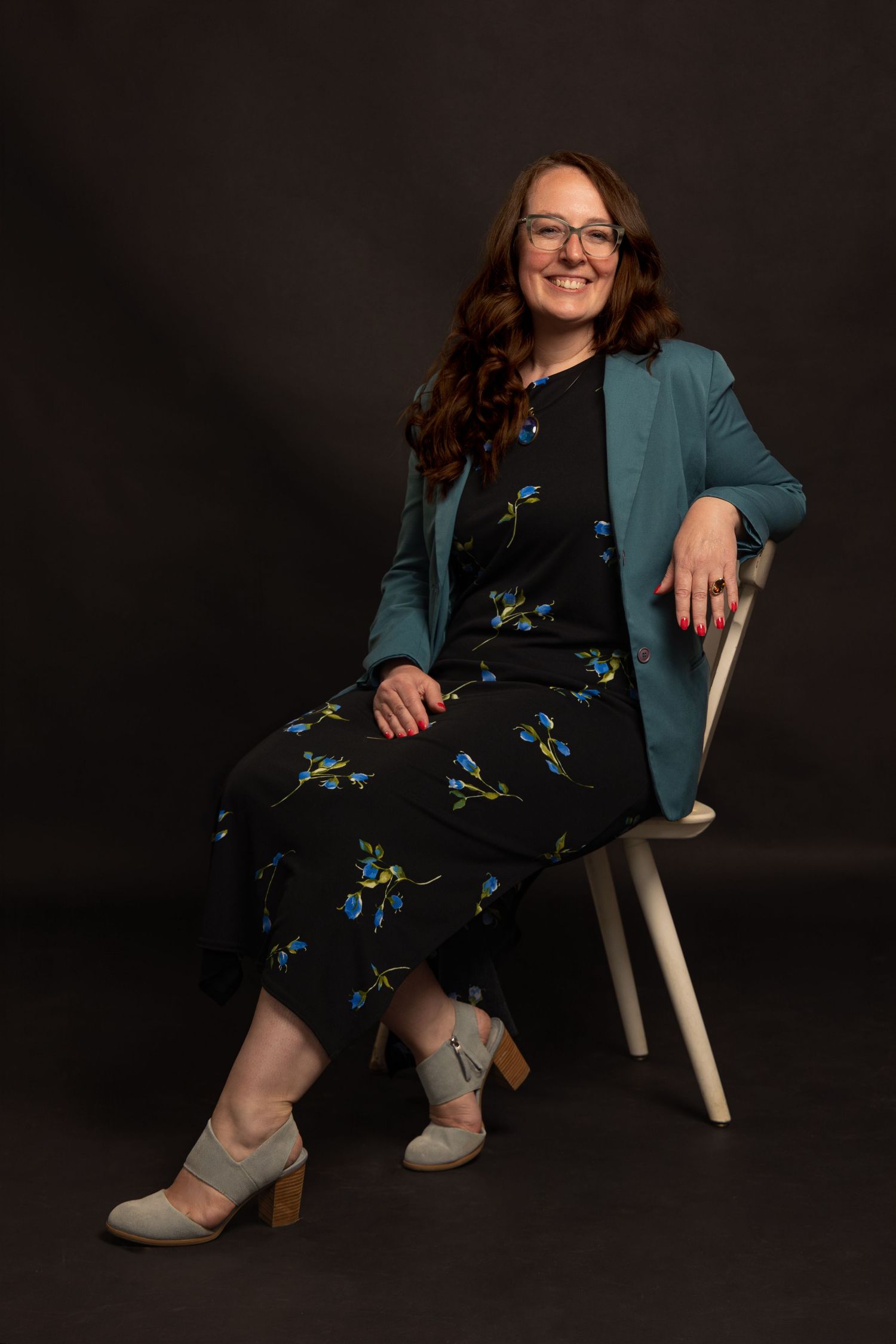 full body portrait of a middle aged brunette woman sitting on a white chair on a black backdrop wearing a blue blazer and dark floral dress