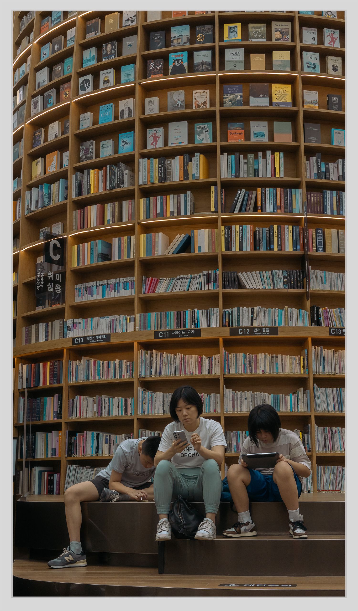 photojournalistic family group mom and 2 kids looking at mobile phone, games while sitting on steps in front of a wall of books in library