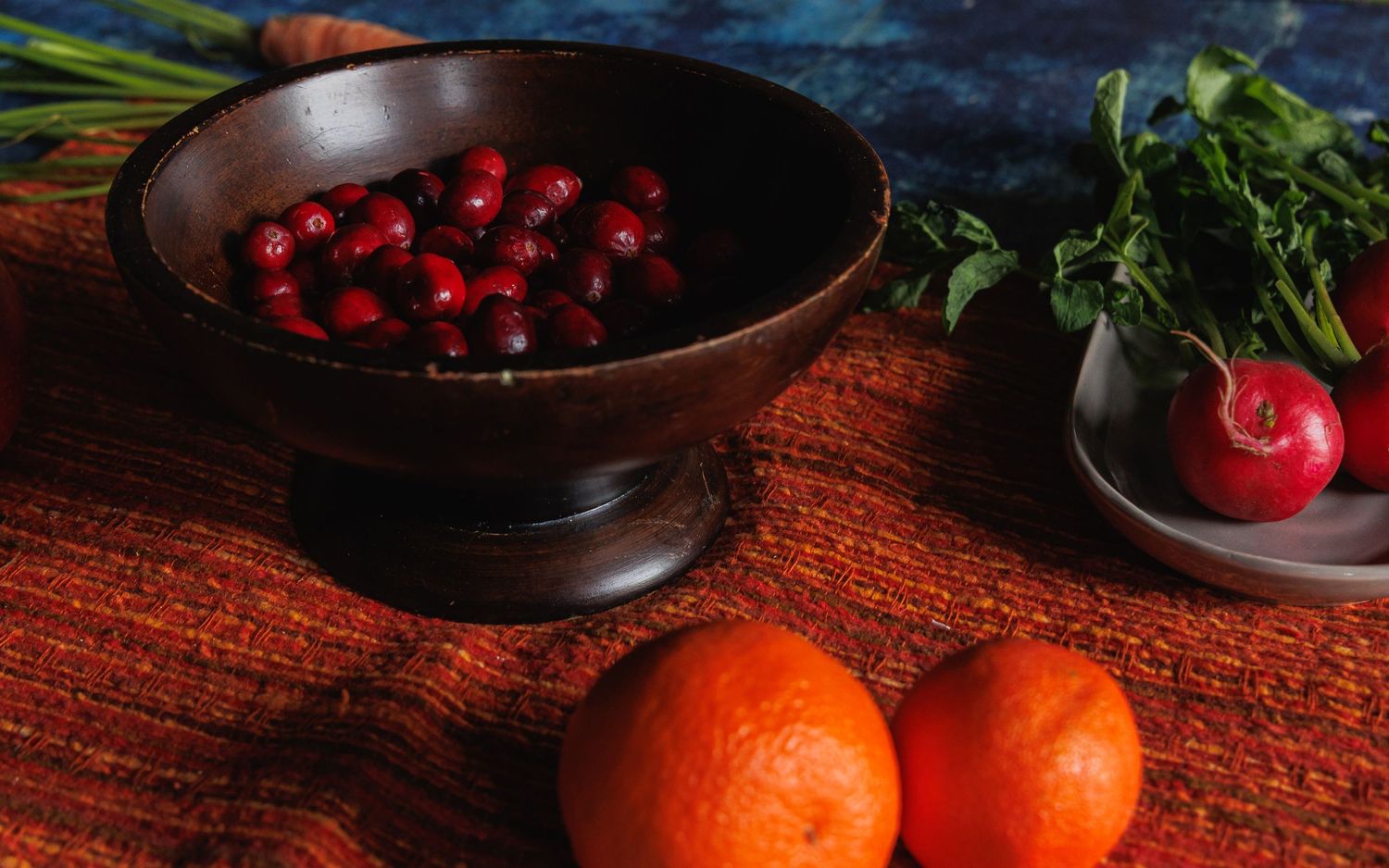 still life image of a table scape with blue and orange table clothes, a wooden bowl of cranberries, grey plate of radishes and 2 oranges