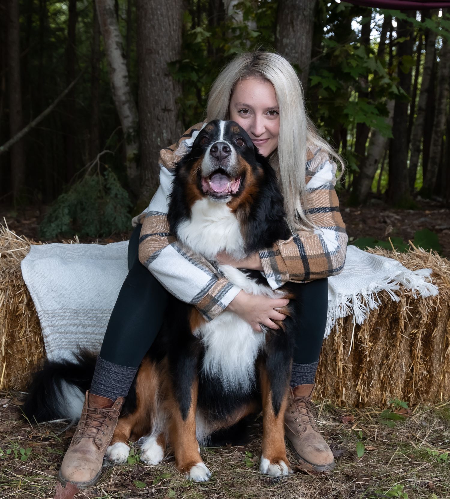 blonde woman hugging her Bernese mountain dog in front of her while she sits on a hay bail in a forest wearing a plaid shirt brown boots