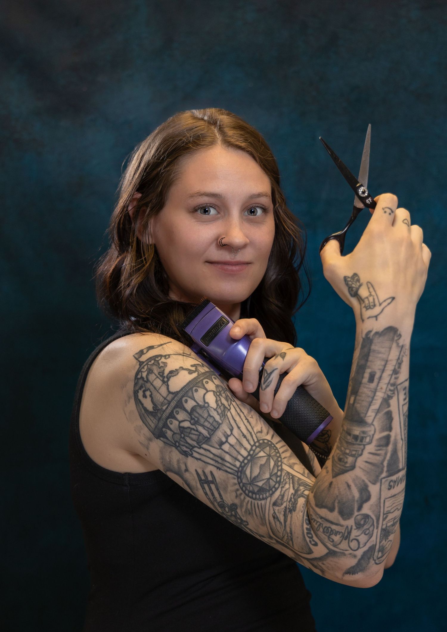 brunette female hairstylist and barber with a full sleeve tattoo flexing her muscles while holding hair clippers and scissors