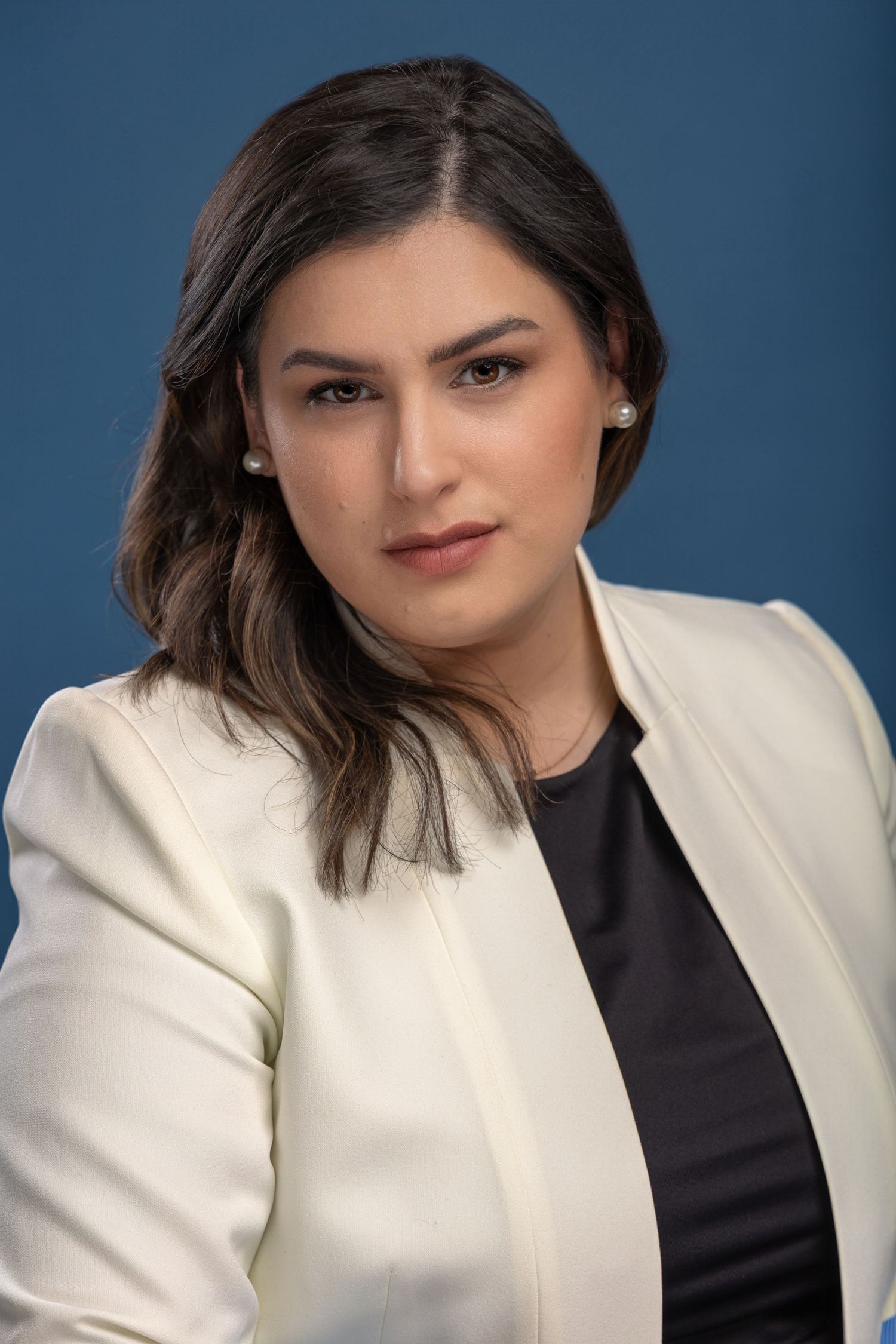 brunette female headshot wearing white blazer and black shirt in front of blue backdrop