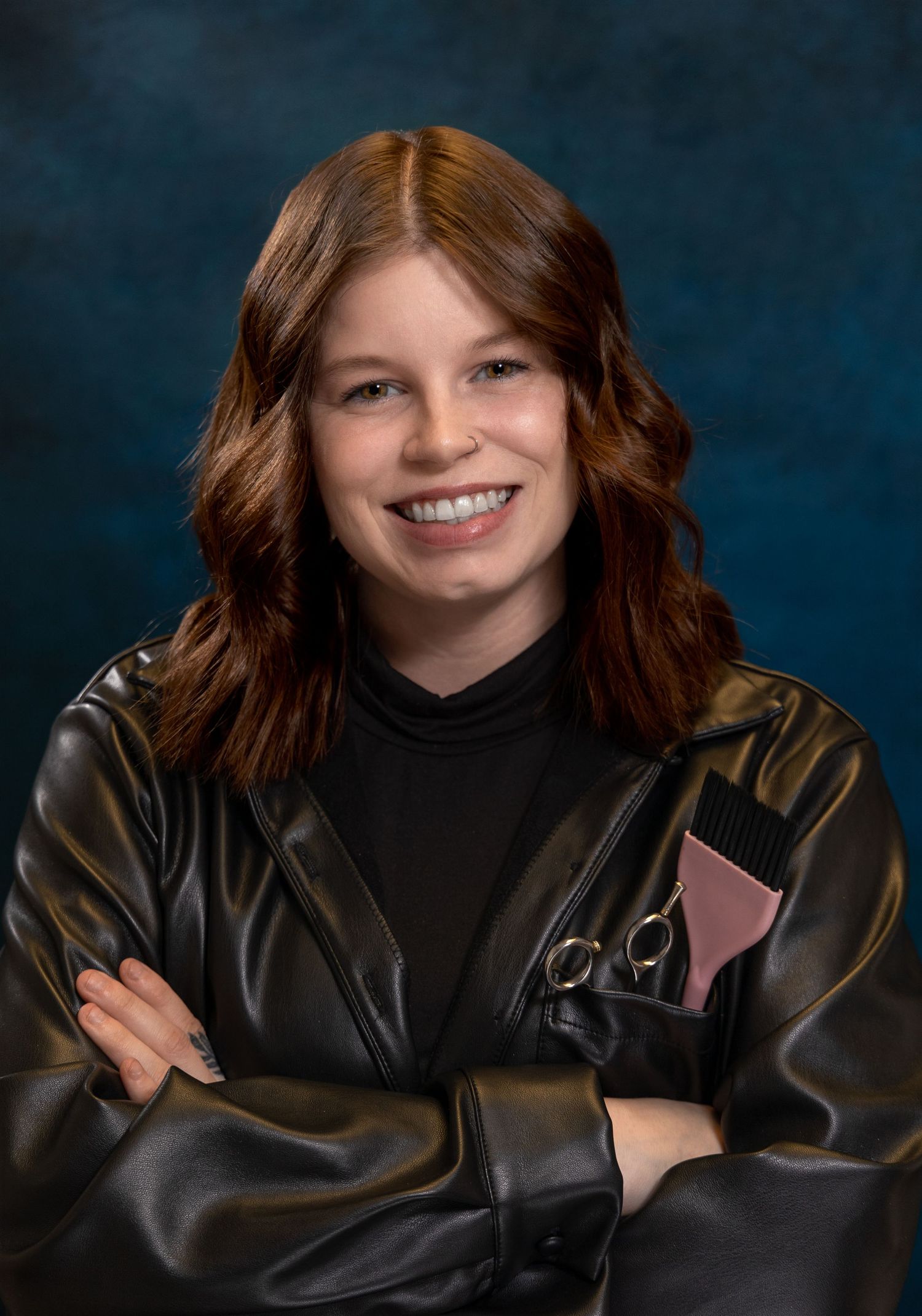 brunette female hairstylist wearing a black leather jacket and shirt with hair tools in her breast pocket