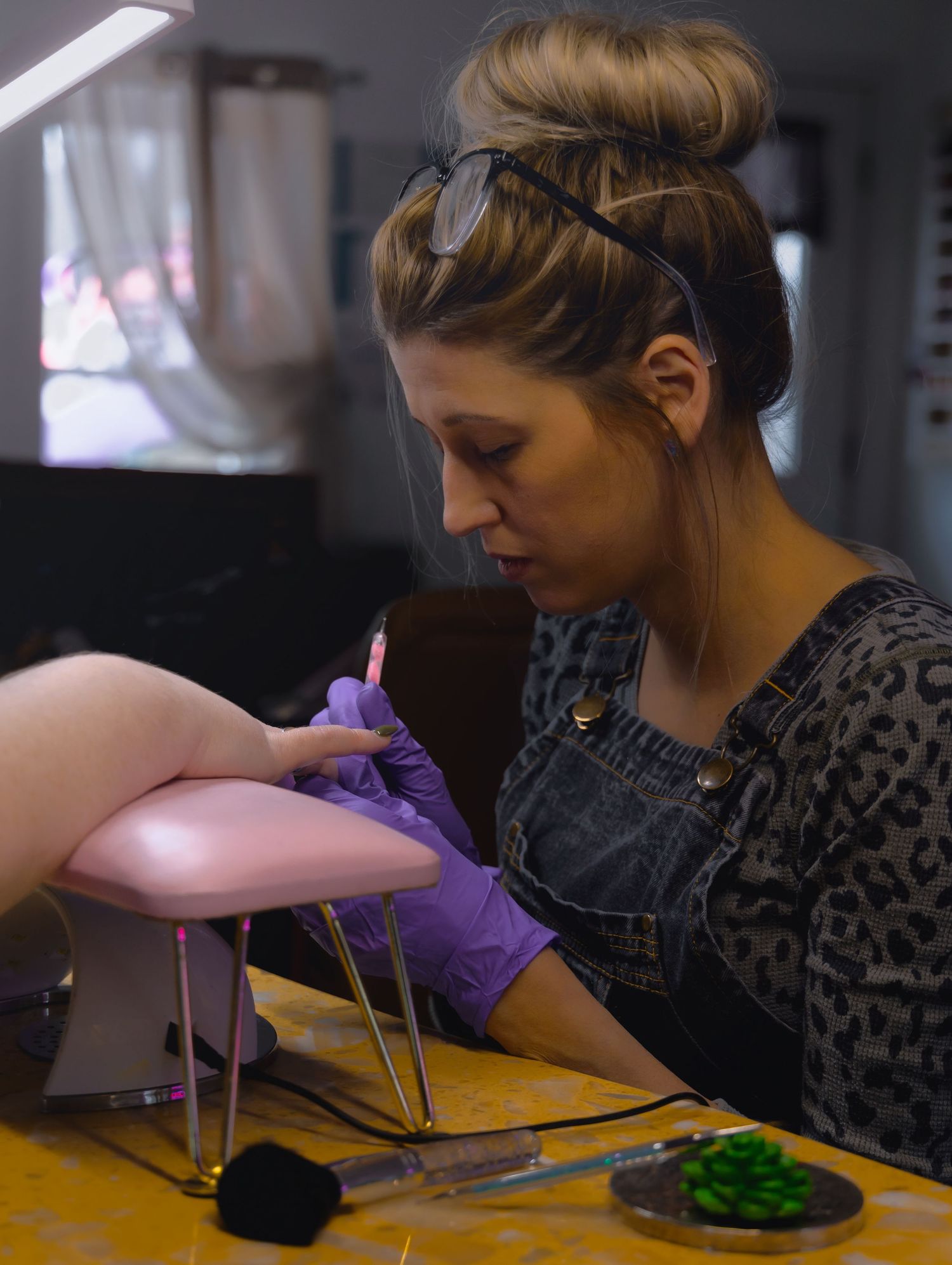 brand image of a blonde female nail tech wearing glasses painting the nails of a client whose hands are on a pink wrist rest