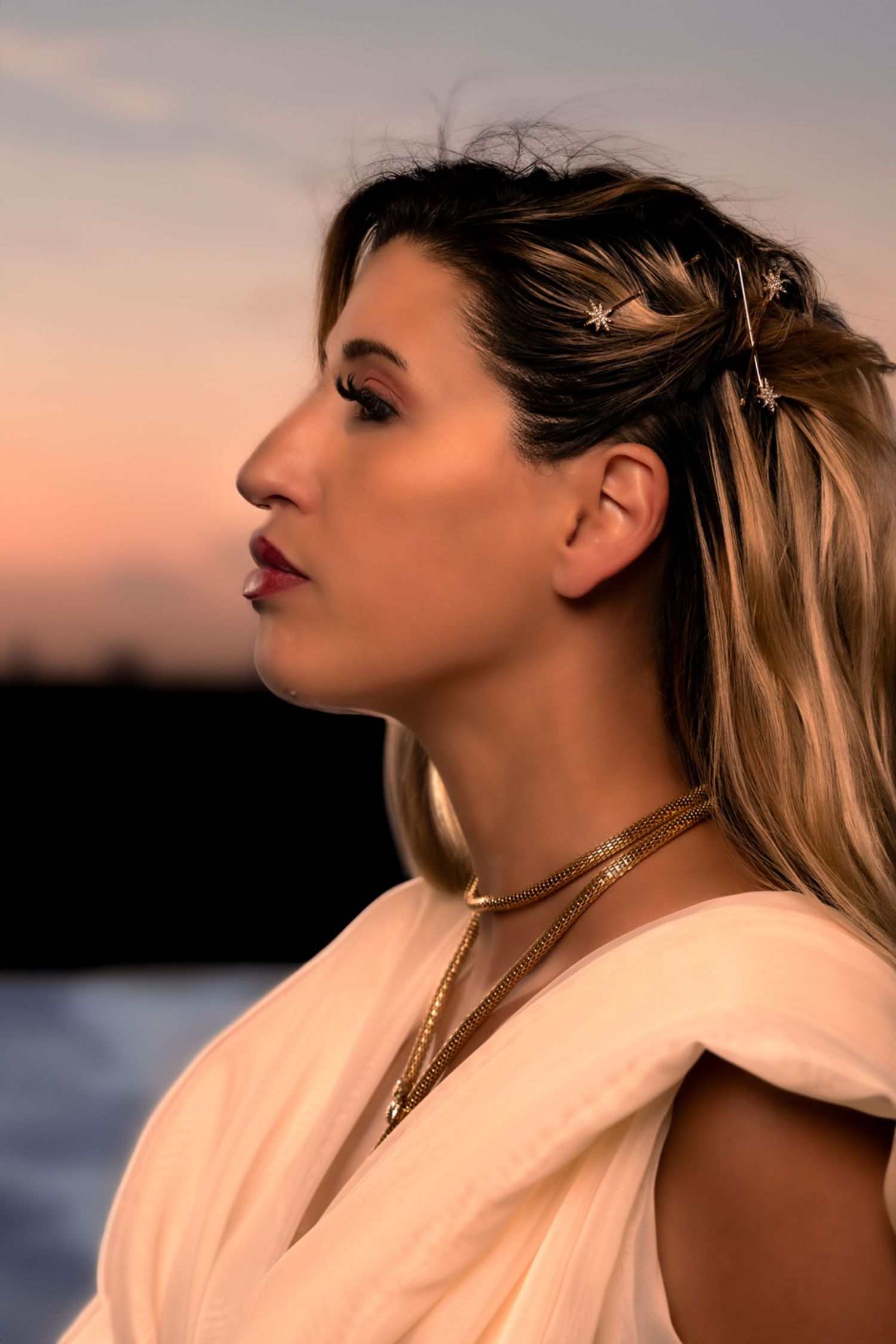 fantasy profile portrait of a blonde woman wearing a white gown and gold necklace looking up river at sunset