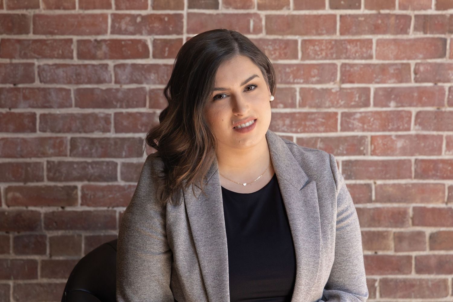 horizontal headshot of a brunette female wearing a black shirt and grey blazer seated in front of a brick wall