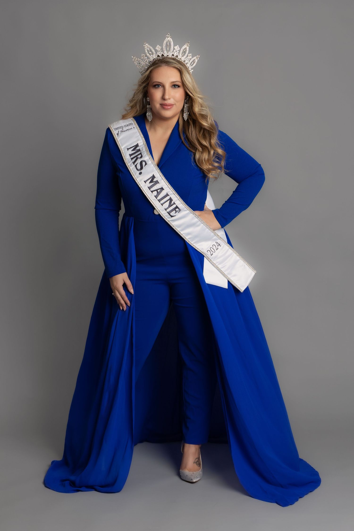 pageant female queen full body portrait wearing crown sash and blue business jumpsuit with a long cape skirt on a grey backdrop