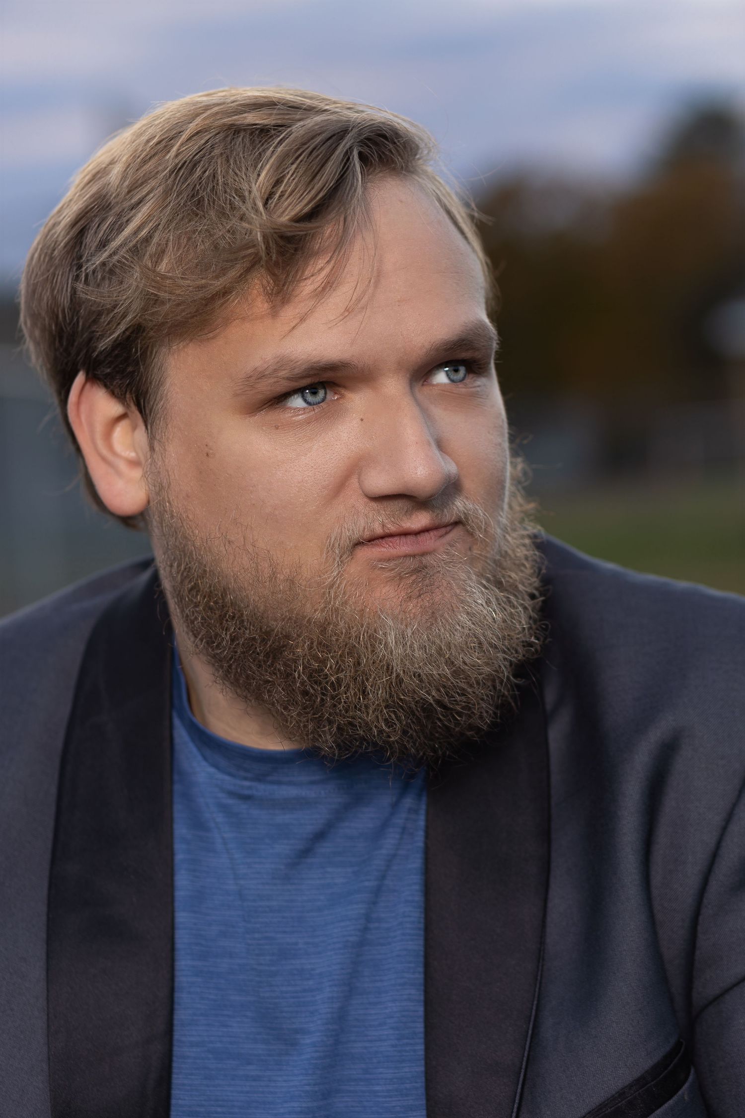 headshot style senior portrait of a blonde male with a bear wearing a grey blazer and blue shirt looking off camera right