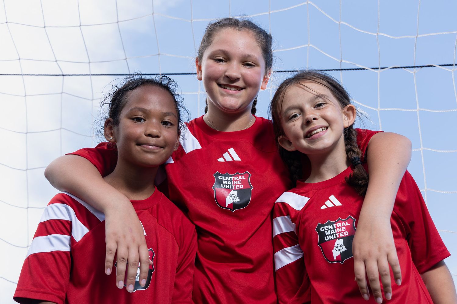 soccer team group shot 3 young girls posed as friends wearing red uniforms standing in a soccer goal smiling