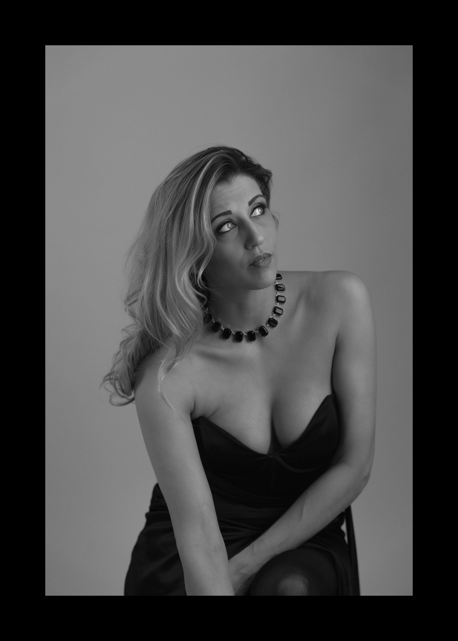 black and white portrait of blonde female seated looking off camera right leaning forward black dress and necklace on white background