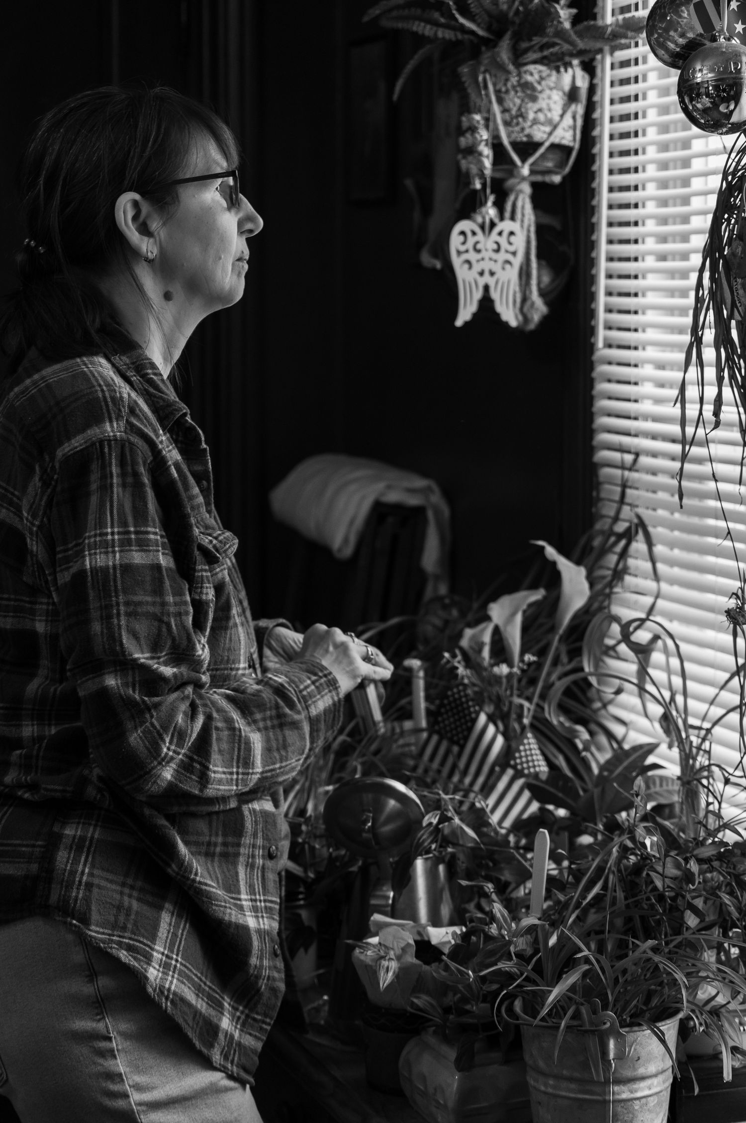 black and white profile image of a middle aged woman looking out her window while tending to a table of plants wearing a plaid shirt