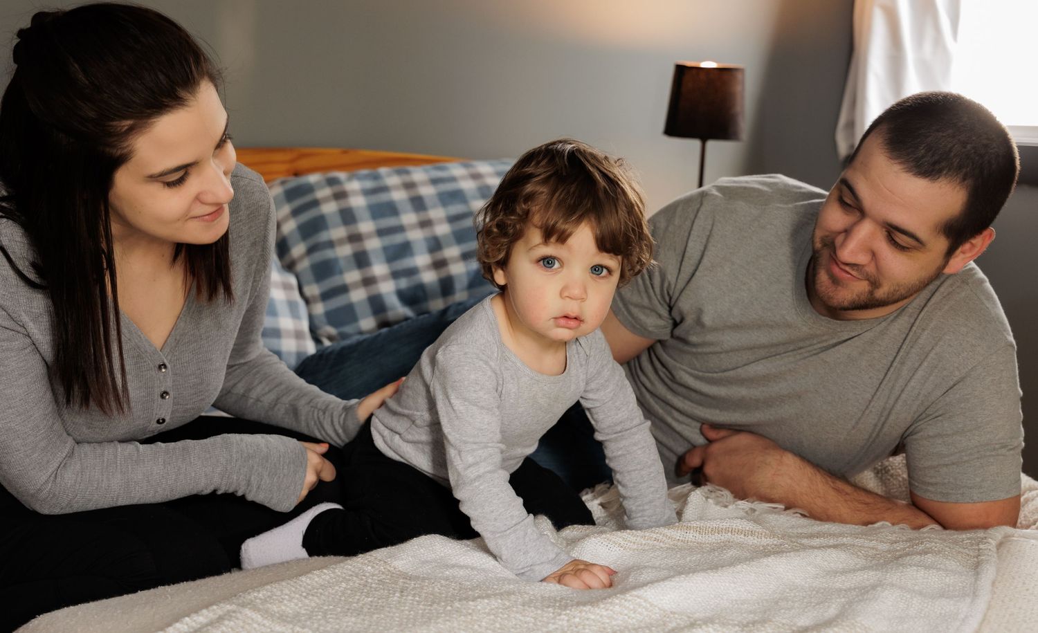 horizontal family image of 2 parents looing at a toddler all wearing grey shirts sitting or laying down on a bed with a white blanket
