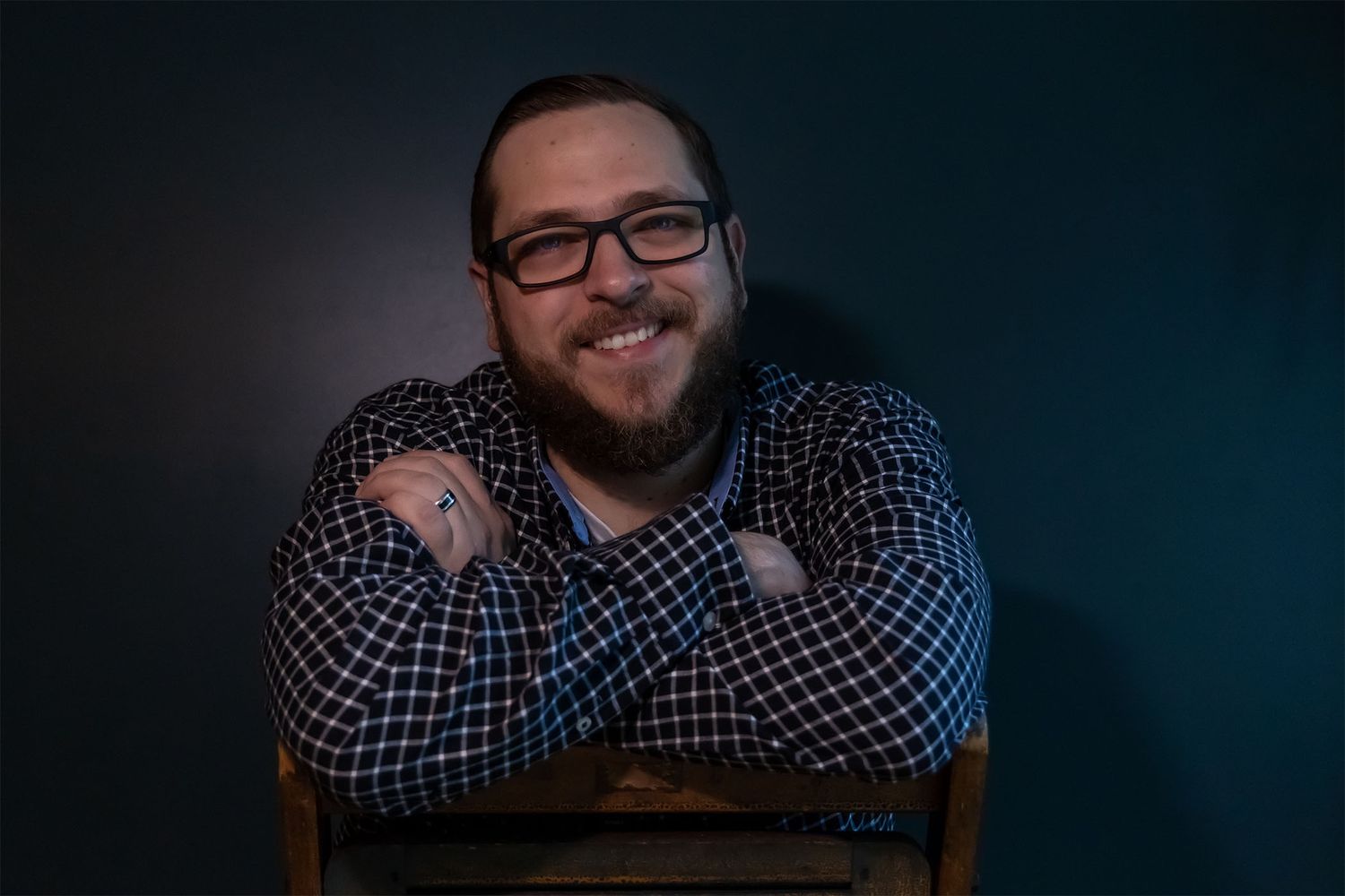horizontal casual seated headshot of a brunette male with his arms crossed, wearing blue plaid and black glasses in front of a blue wall