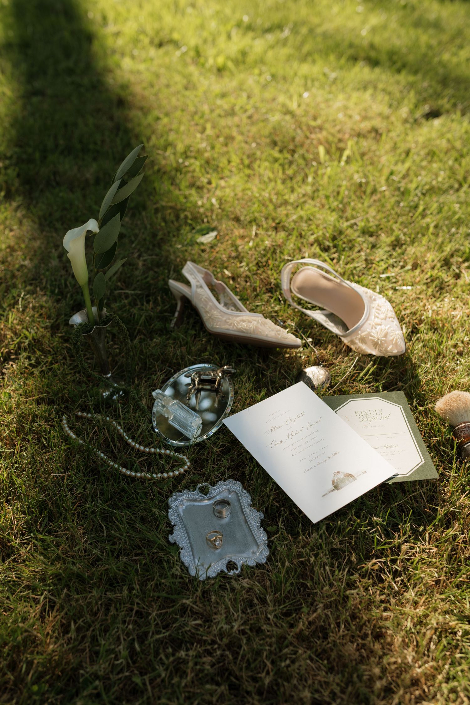Wedding flat lay with lace heels, pearl necklace, rings, perfume, and invitation on green grass.