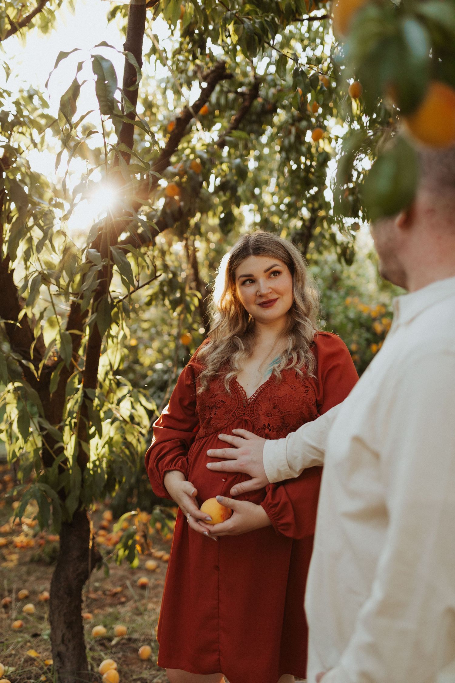 Pregnant woman in red dress cradles baby bump while partner touches belly in golden sunlit orchard.