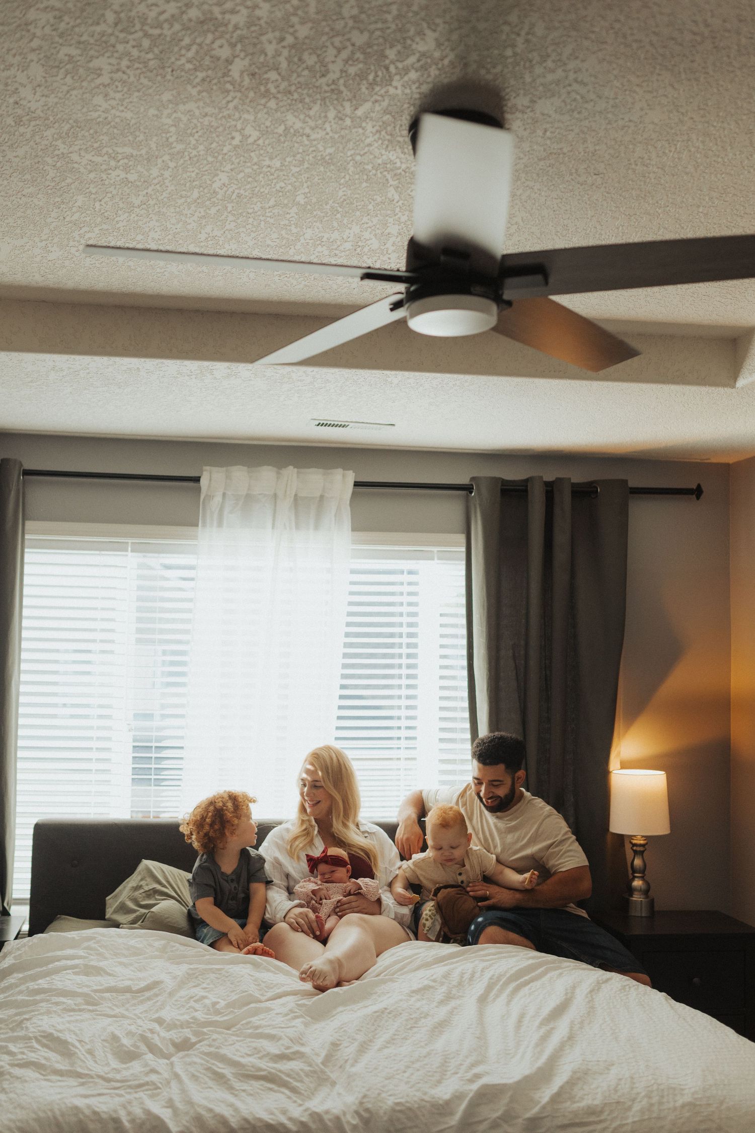 A family of four sits together on a large white bed near a window with a ceiling fan above.