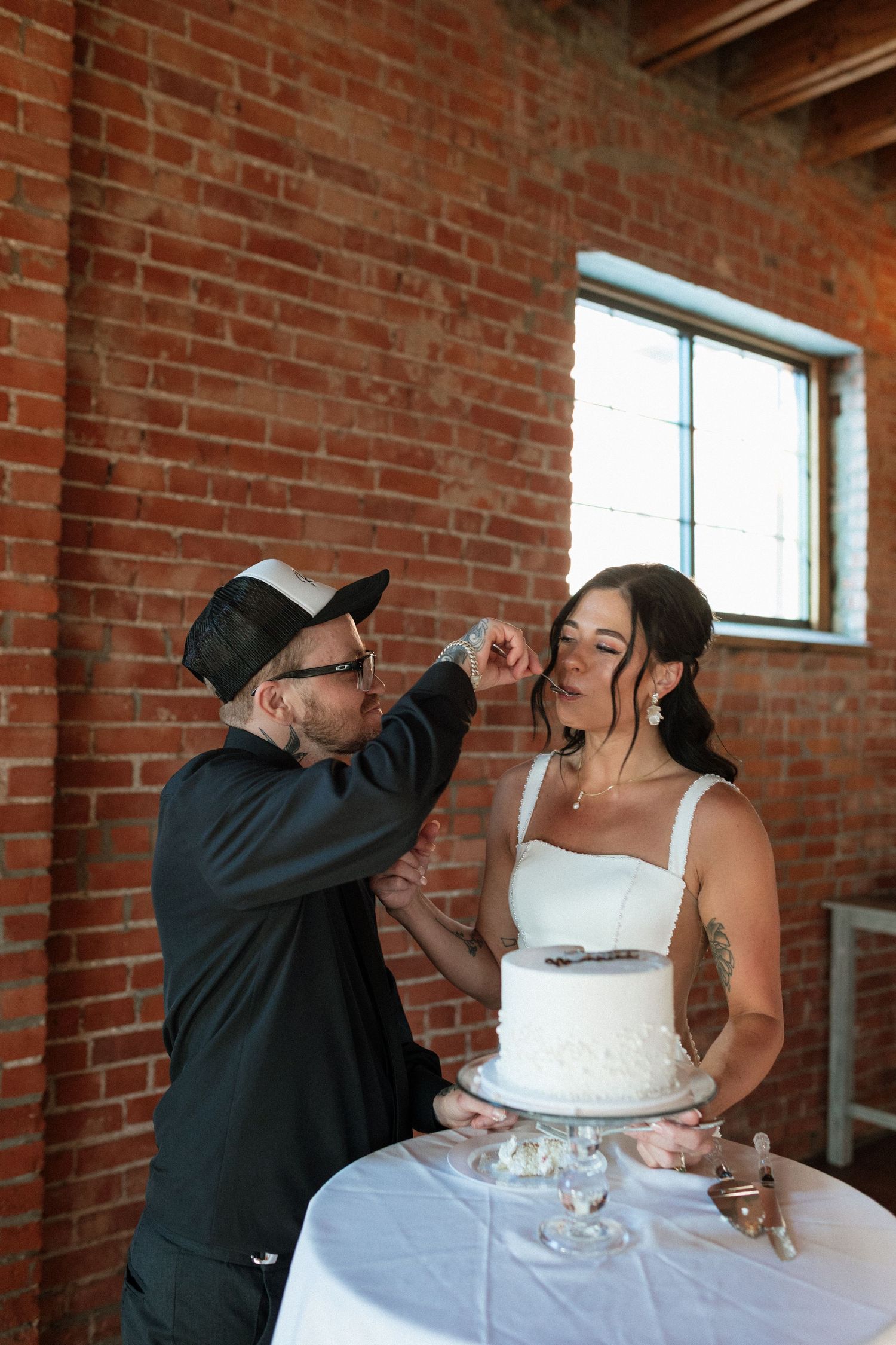 A groom in a black cap feeds wedding cake to his laughing bride in front of a rustic brick wall.
