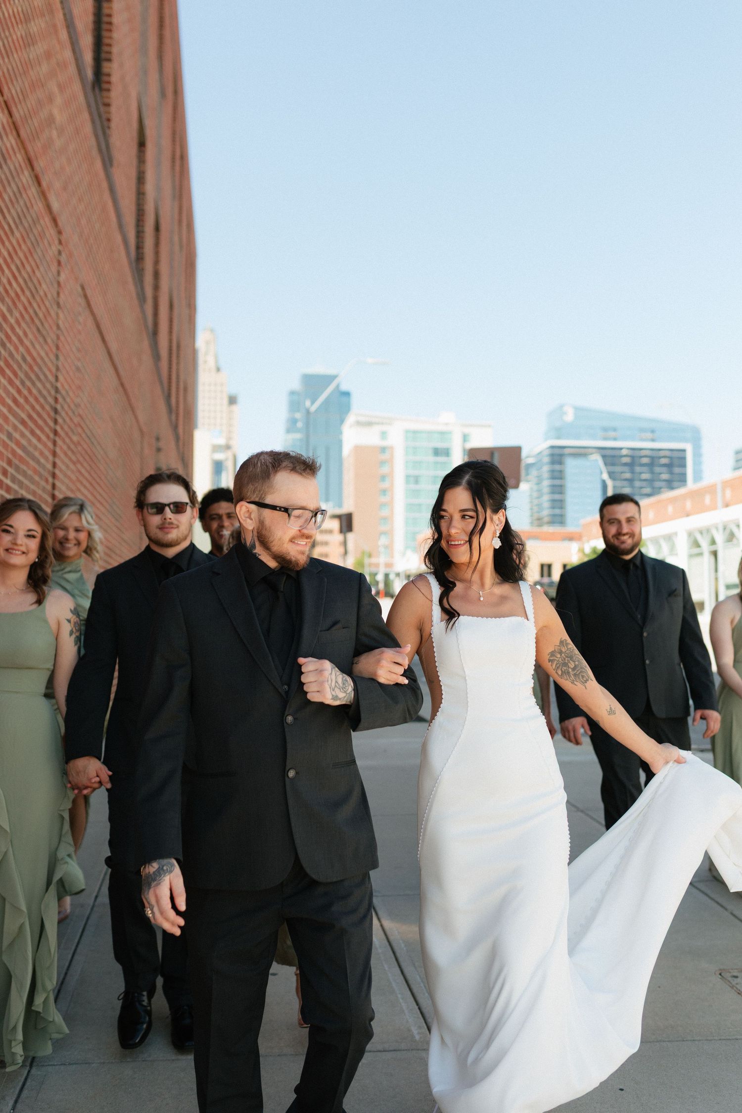 Bride and groom lead wedding party walking outdoors along brick building with city skyline backdrop.