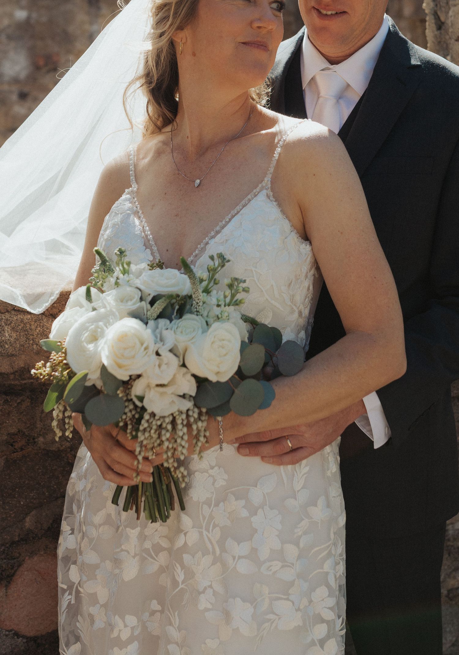 Close-up of bride holding white rose and eucalyptus bouquet with groom embracing her from behind.