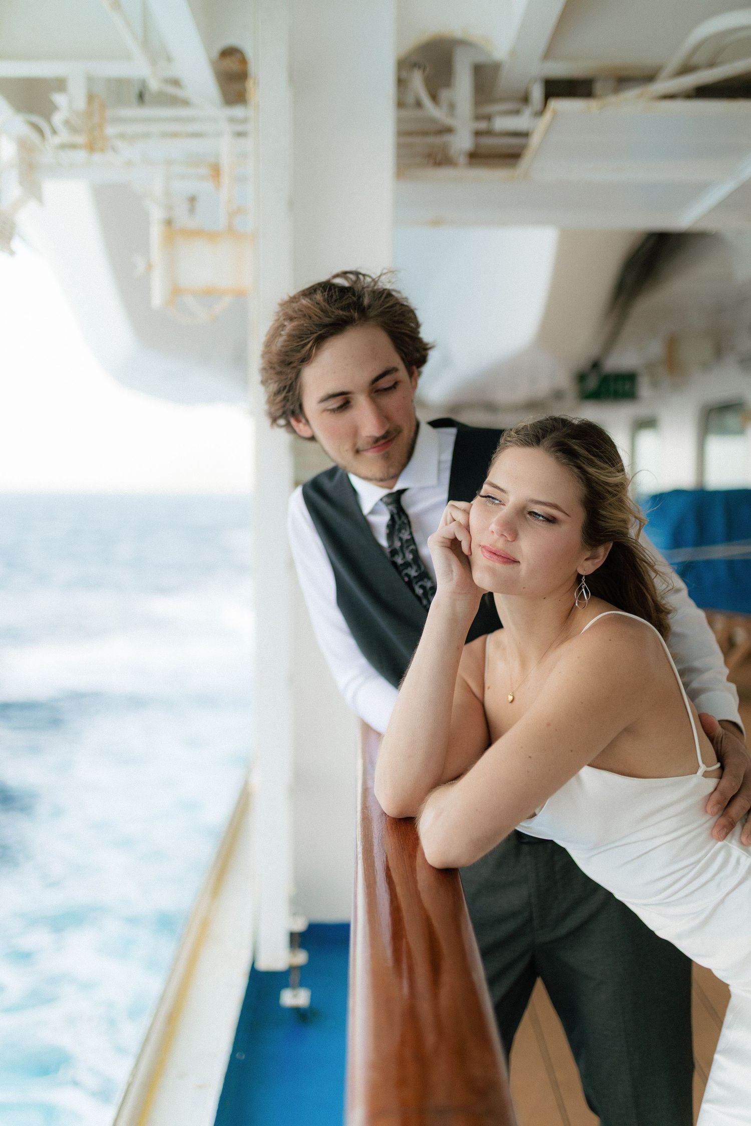 Couple leans on cruise ship railing together, gazing at the blue ocean in formal attire.