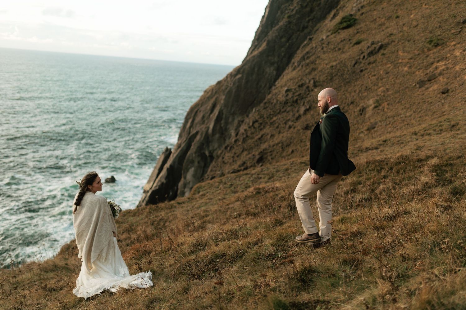 Bride and groom standing on a rugged coastal hillside with dramatic ocean cliffs.