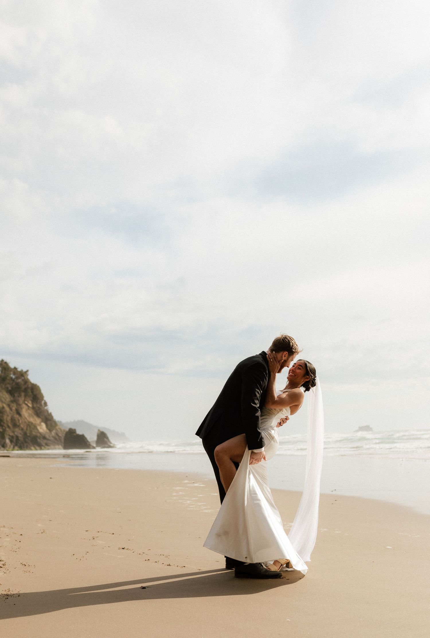 Groom dips bride for a romantic kiss on sandy beach with ocean waves and rocky cliffs in background.