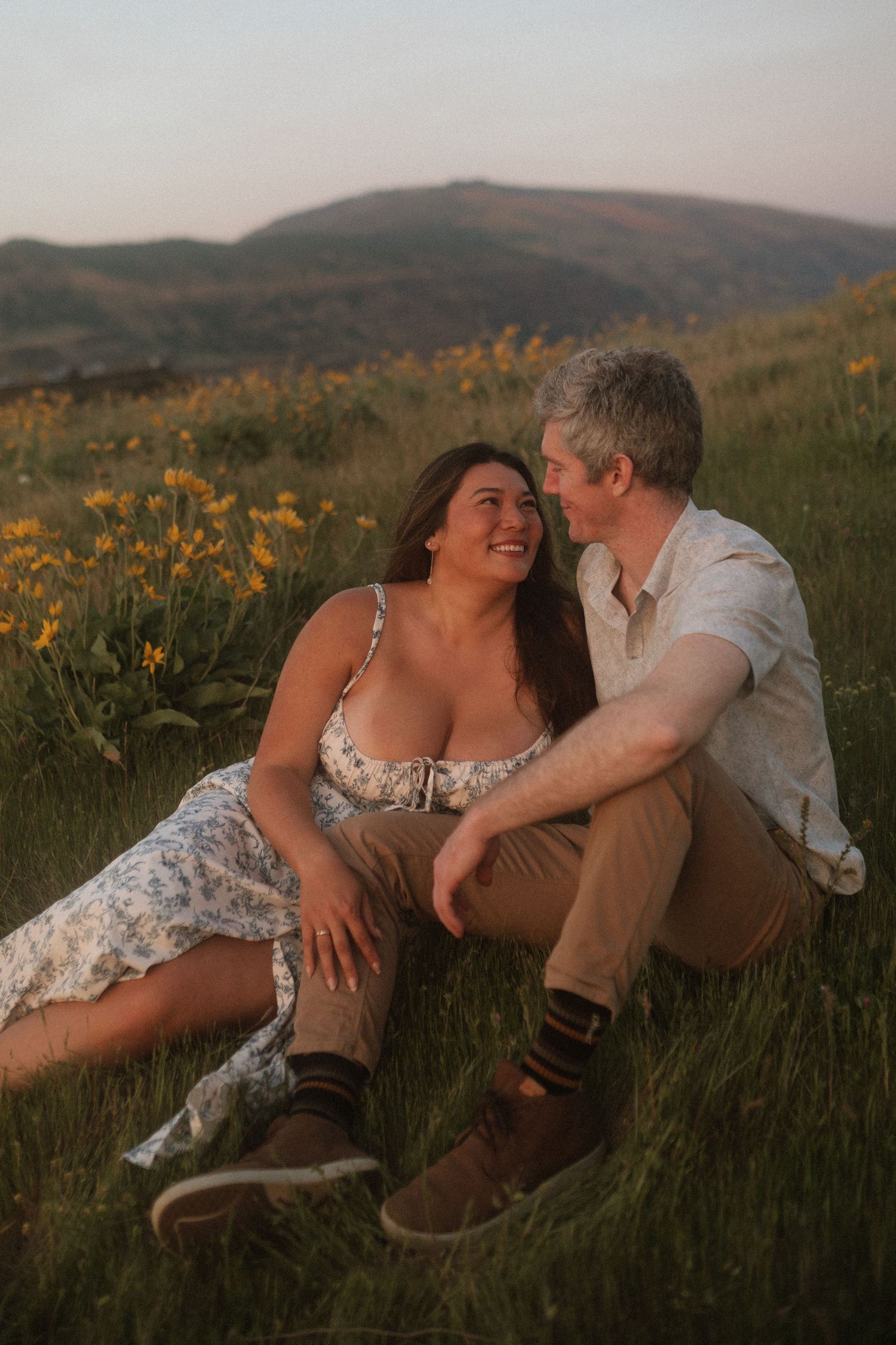 Romantic couple sitting together in a flower-filled meadow at golden hour, smiling and gazing at each other.