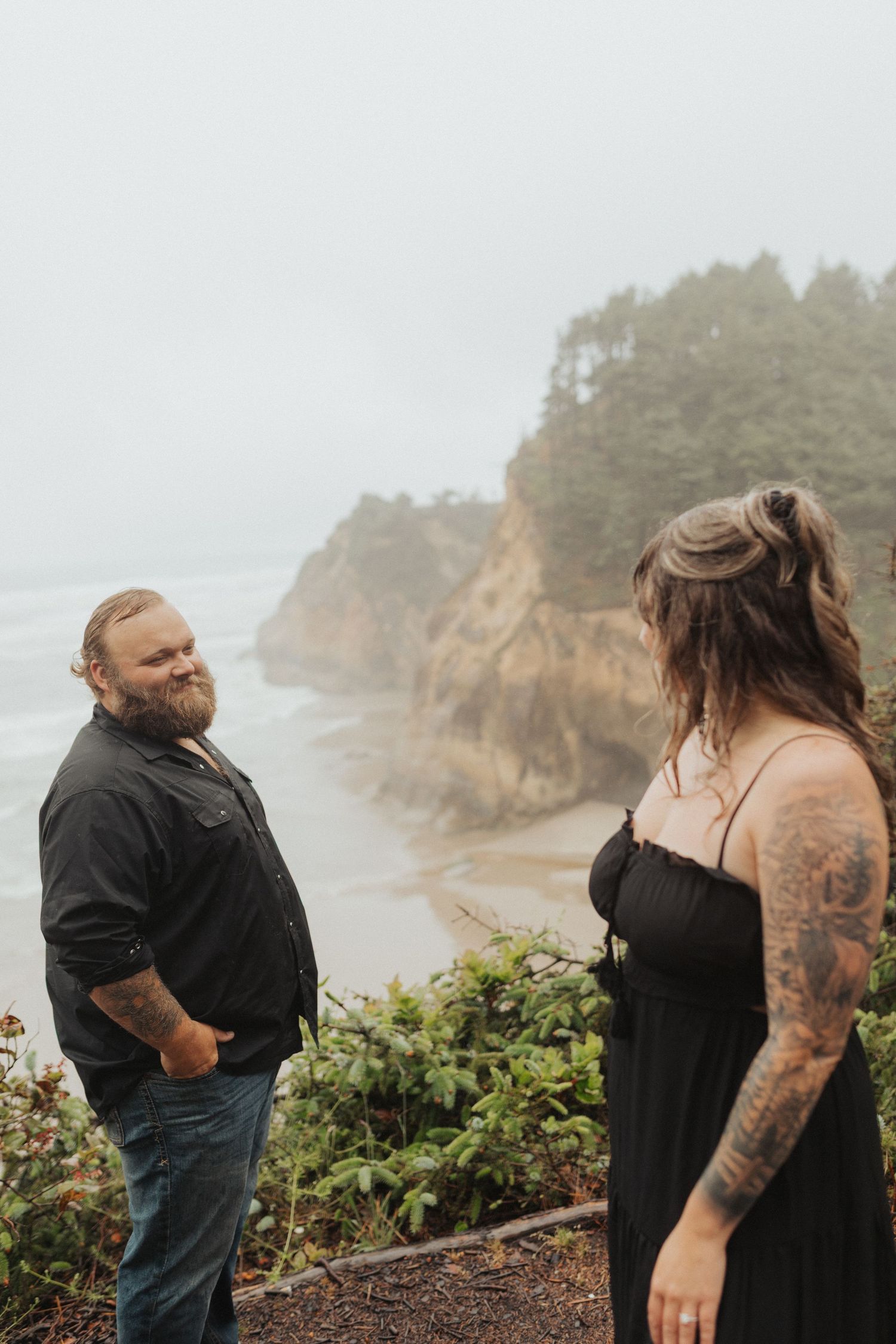 Tattooed couple in black outfits share a moment on foggy Oregon coast overlook with beach below.
