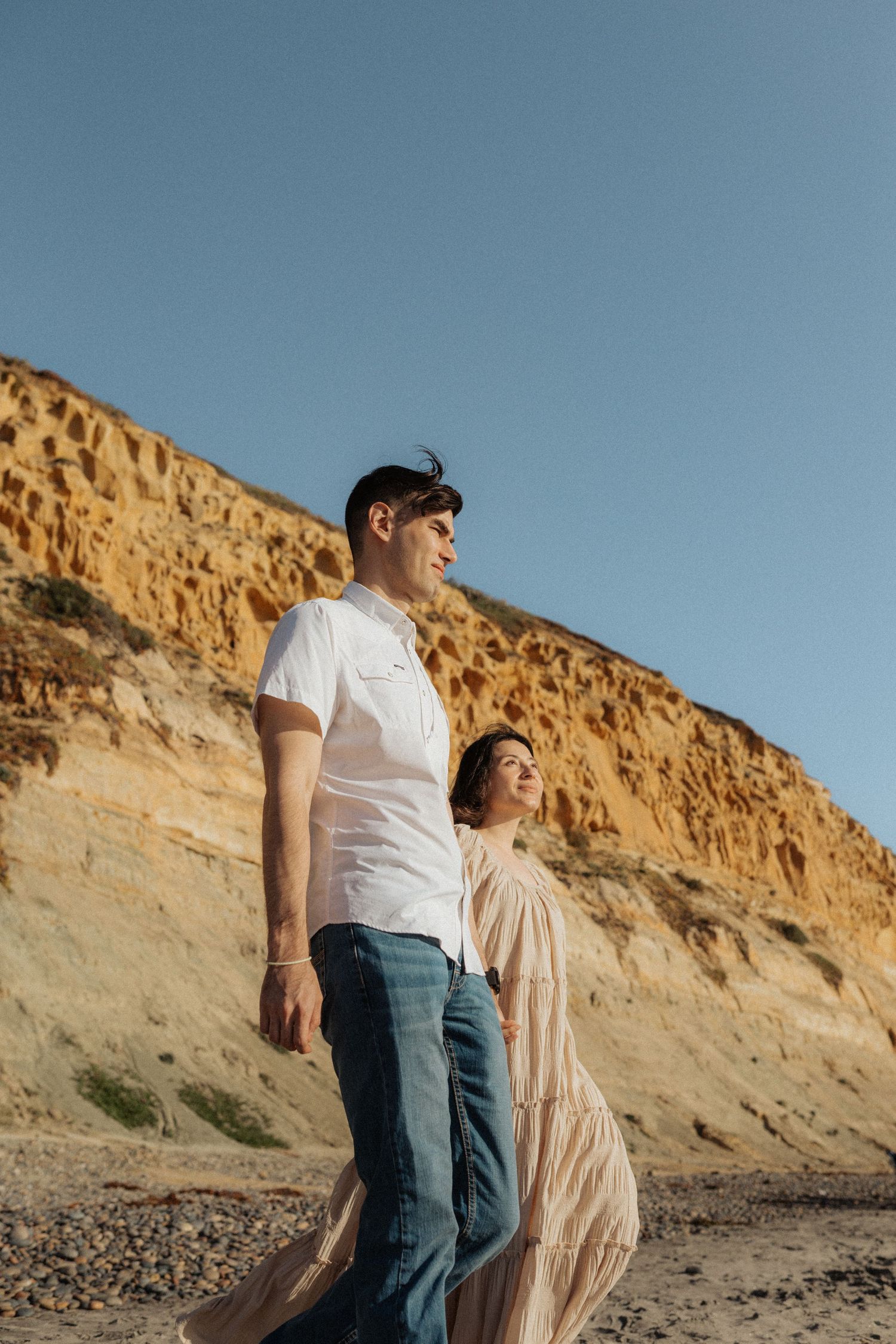 Couple standing together on pebble beach with golden cliffs, man in white tee and jeans, woman in flowing beige dress.