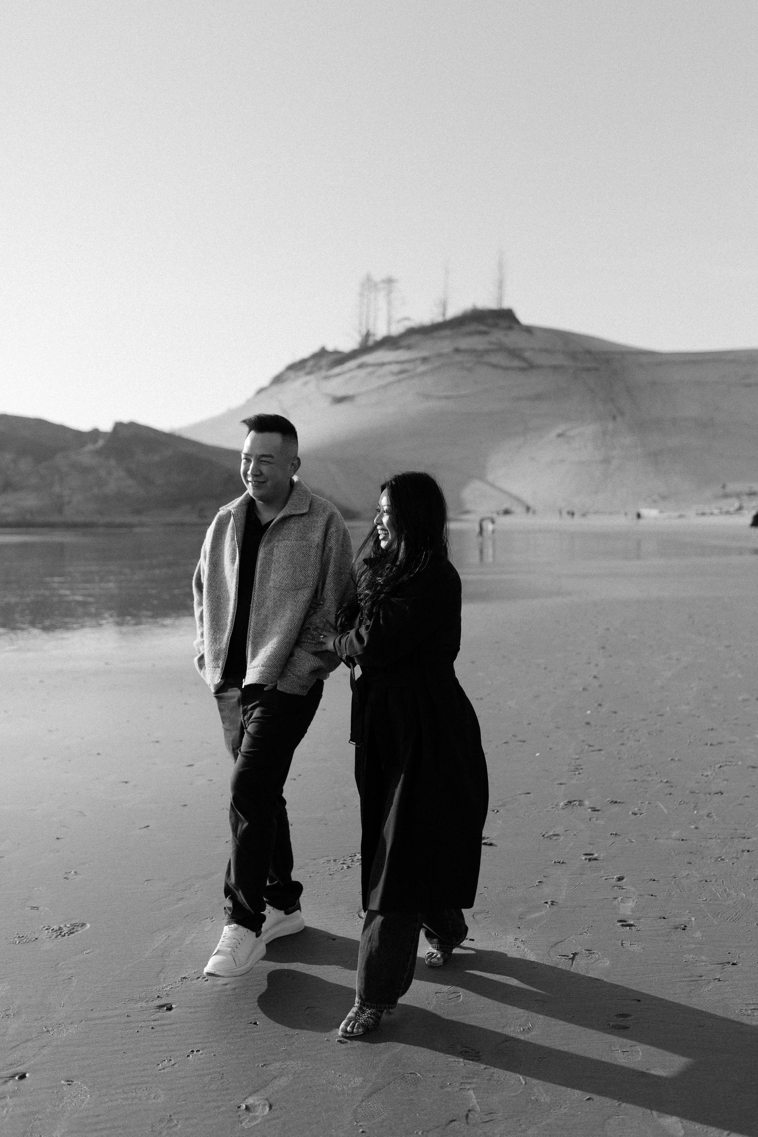 Black and white photo of couple holding hands walking on beach with sand dunes in background.