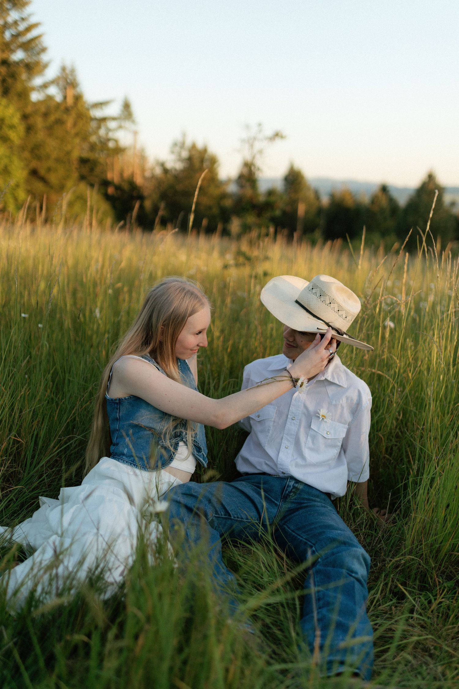 Young woman playfully adjusting cowboy hat on man while sitting together in sunlit green meadow.