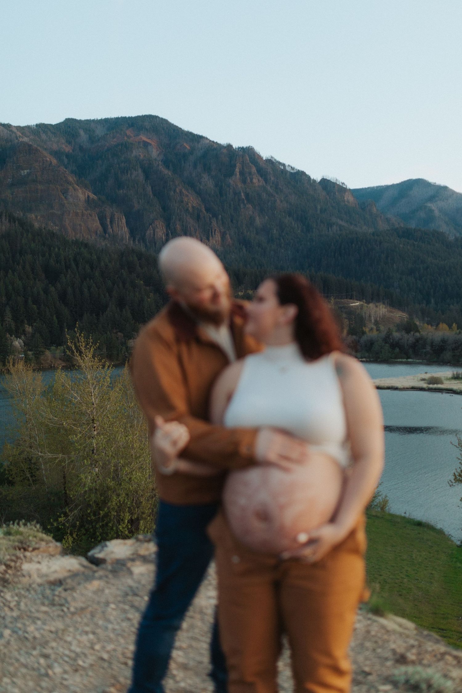 Soft focus image of pregnant couple embracing with a scenic mountain lake and clear sky in background.