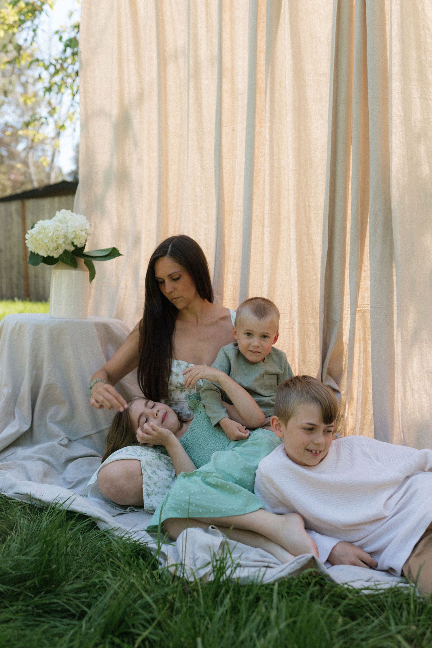 A laughing mother enjoys a playful moment with her three children on a blanket in a garden.