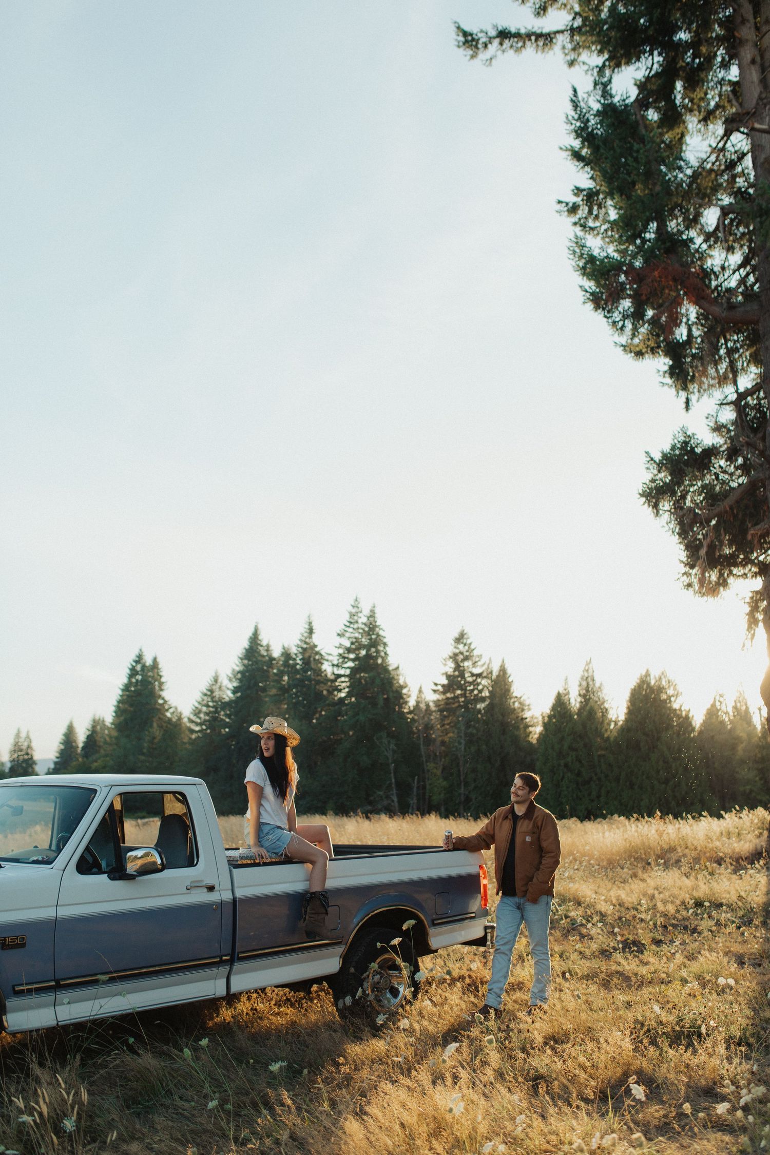 A woman in a cowboy hat sits on a vintage pickup truck while a man stands beside her in a golden field.