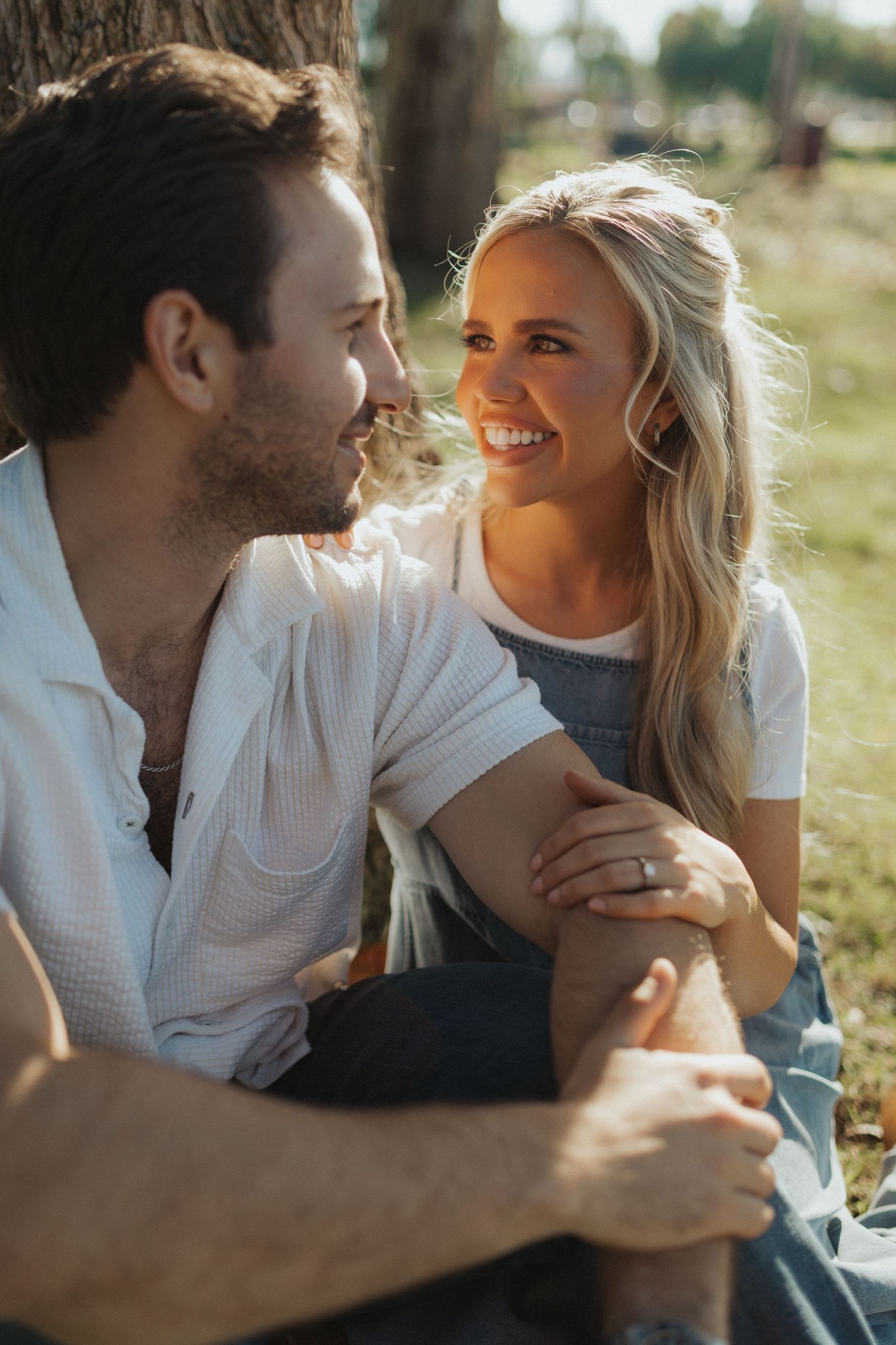 A smiling blonde woman and bearded man sit together against a tree in golden sunlight.