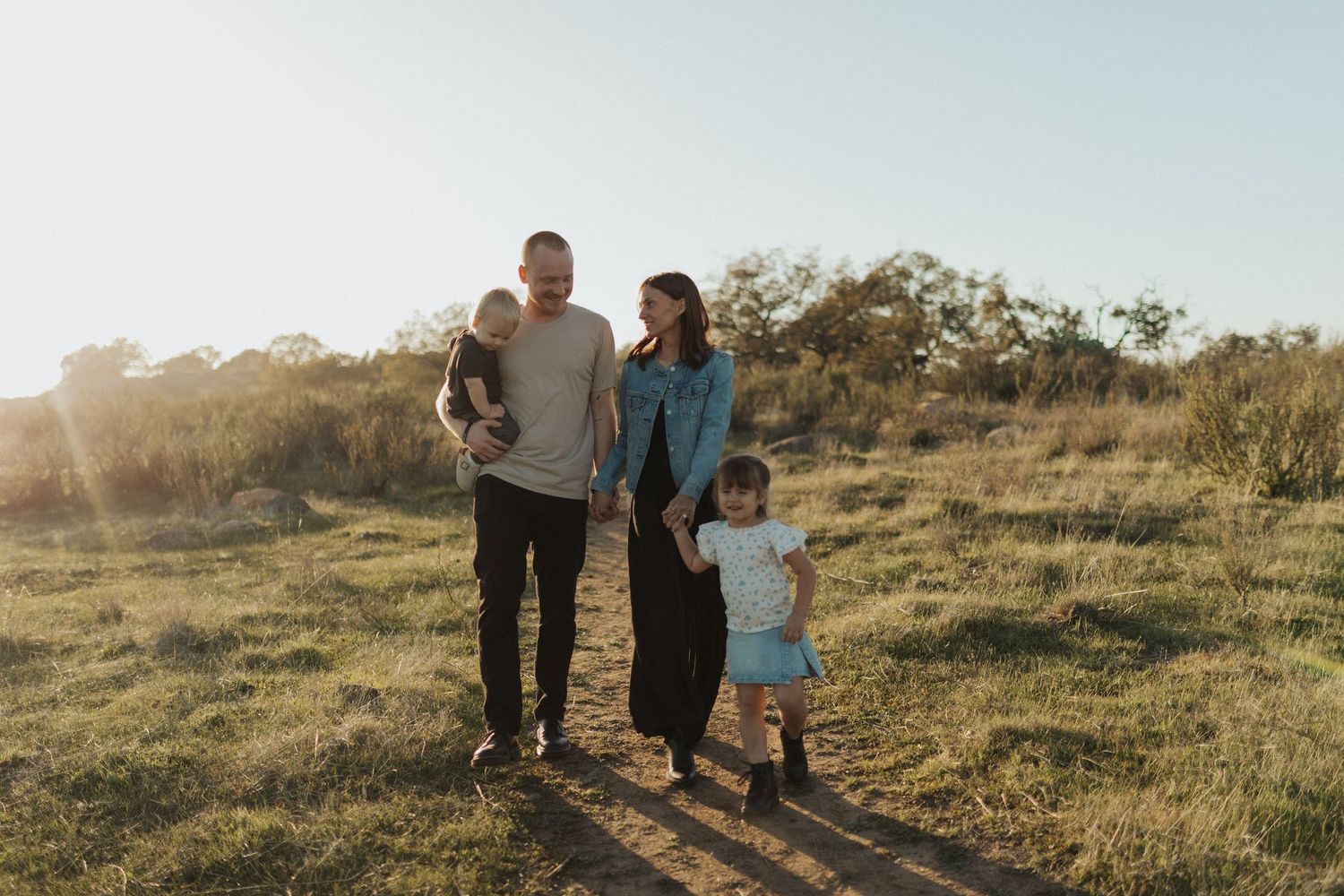Happy family of four walking on sunny grass field path, father holding baby, mother with toddler.